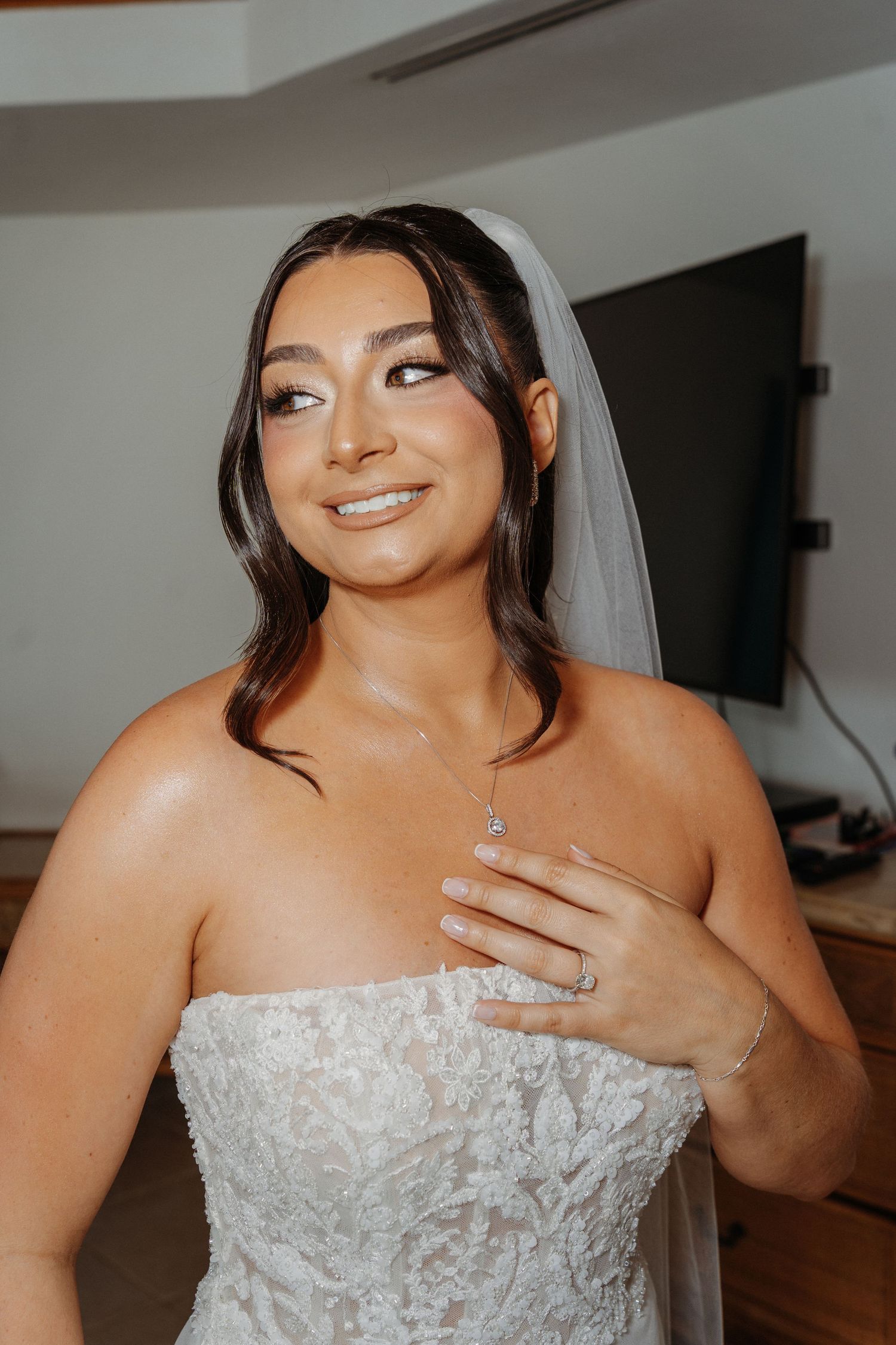 Smiling bride in strapless lace wedding gown and veil, touching her necklace, with elegant bridal makeup.