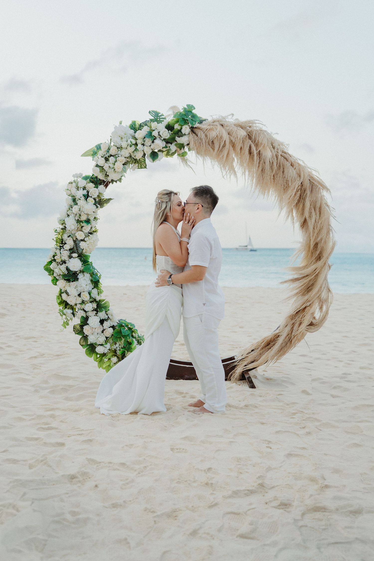 Romantic beach wedding ceremony with floral arch and white decor on sandy shore at sunset.