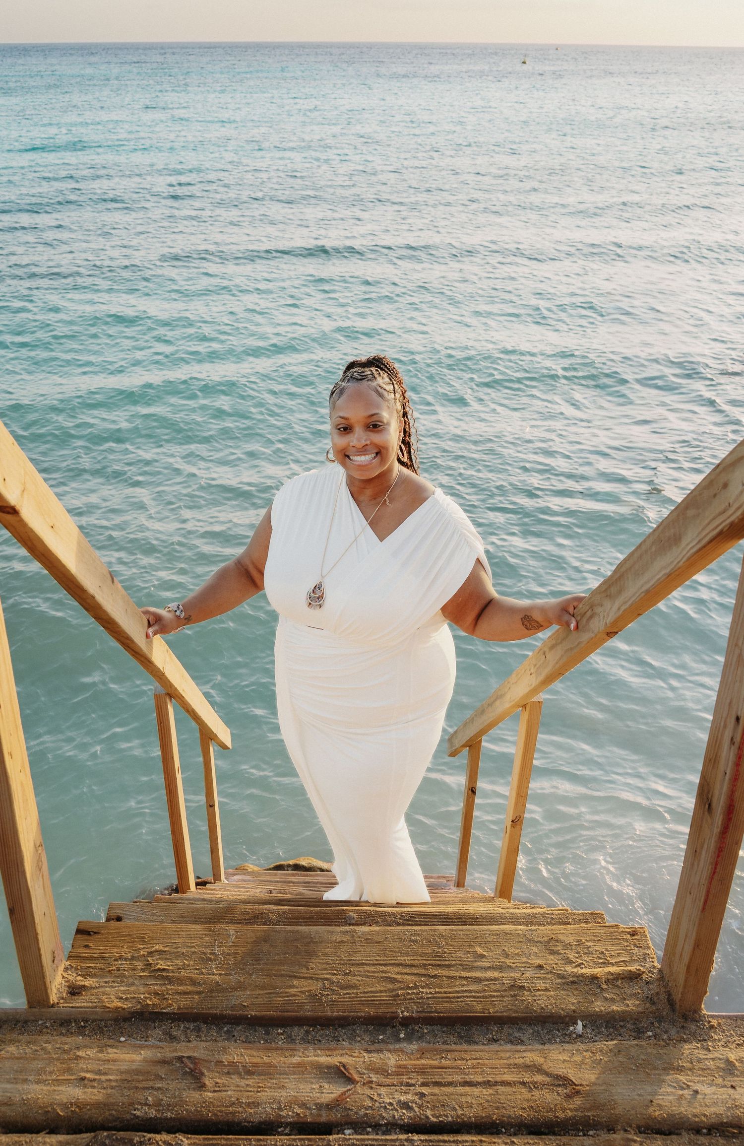 Someone in white stands on wooden steps leading to the beach while smiling warmly at the camera.