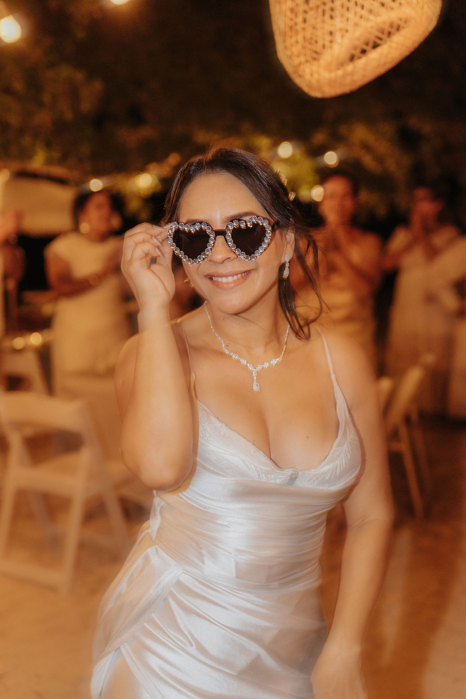 A person in a white satin dress poses playfully wearing heart-shaped sunglasses at an evening event.
