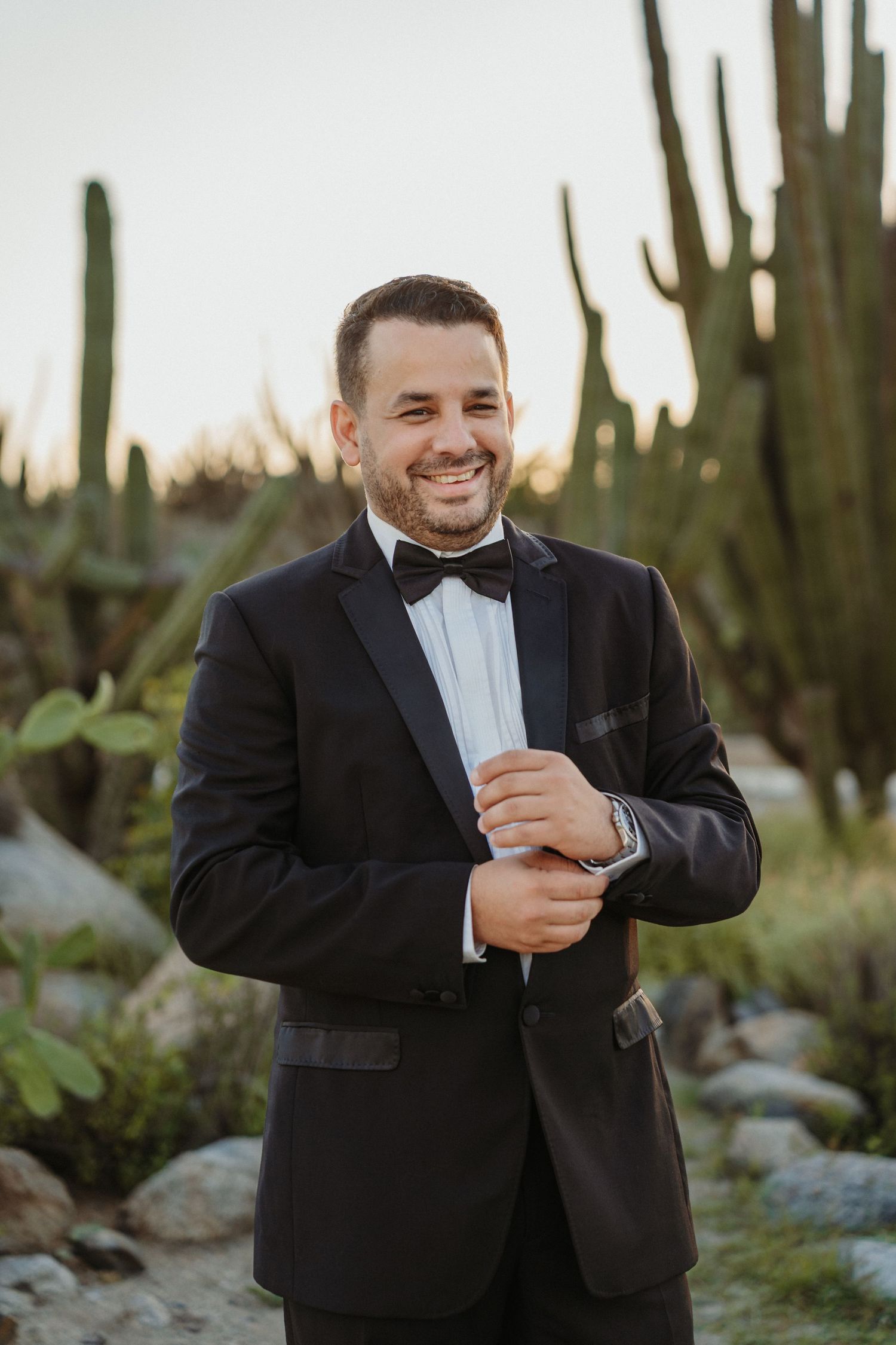 A person in a black tuxedo laughs while holding a white paper in a desert landscape with saguaro cacti.