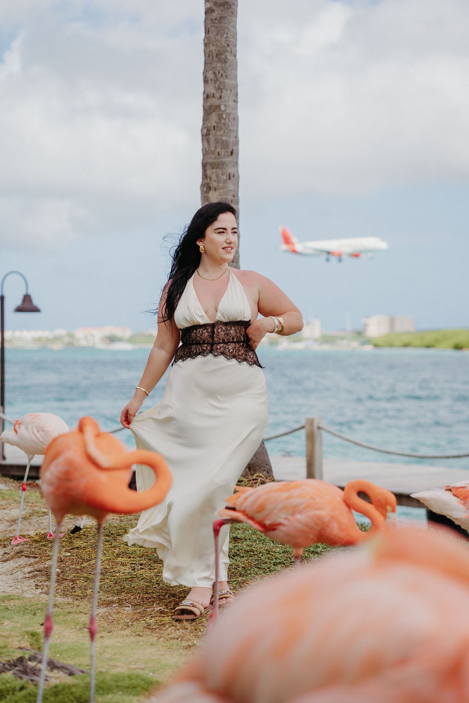 A white maxi dress with lace detail stands among pink flamingo decorations near the ocean as a plane flies overhead.