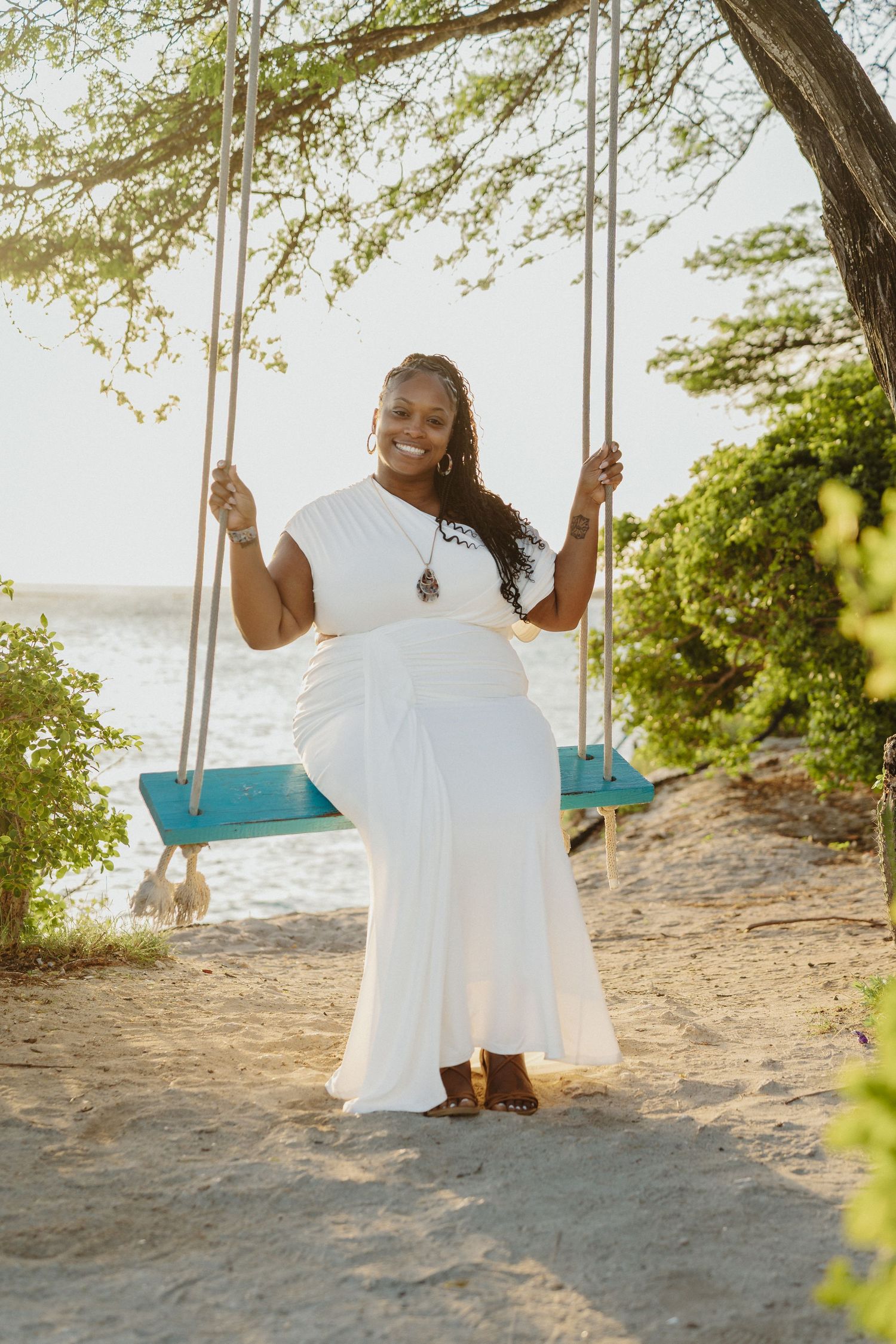 A person in a flowing white dress sits on a wooden swing at a sunny beach with ocean waves visible in the background.