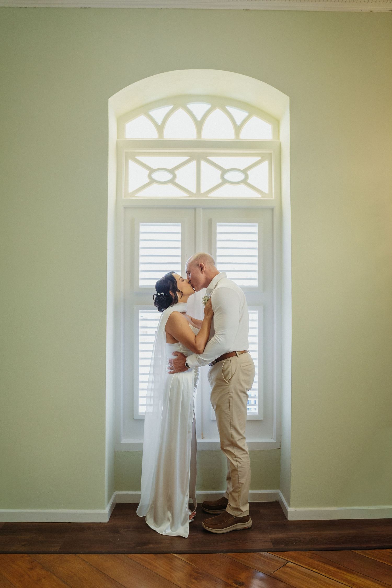 A couple shares a romantic kiss in front of an elegant arched window with white shutters and hardwood floors.