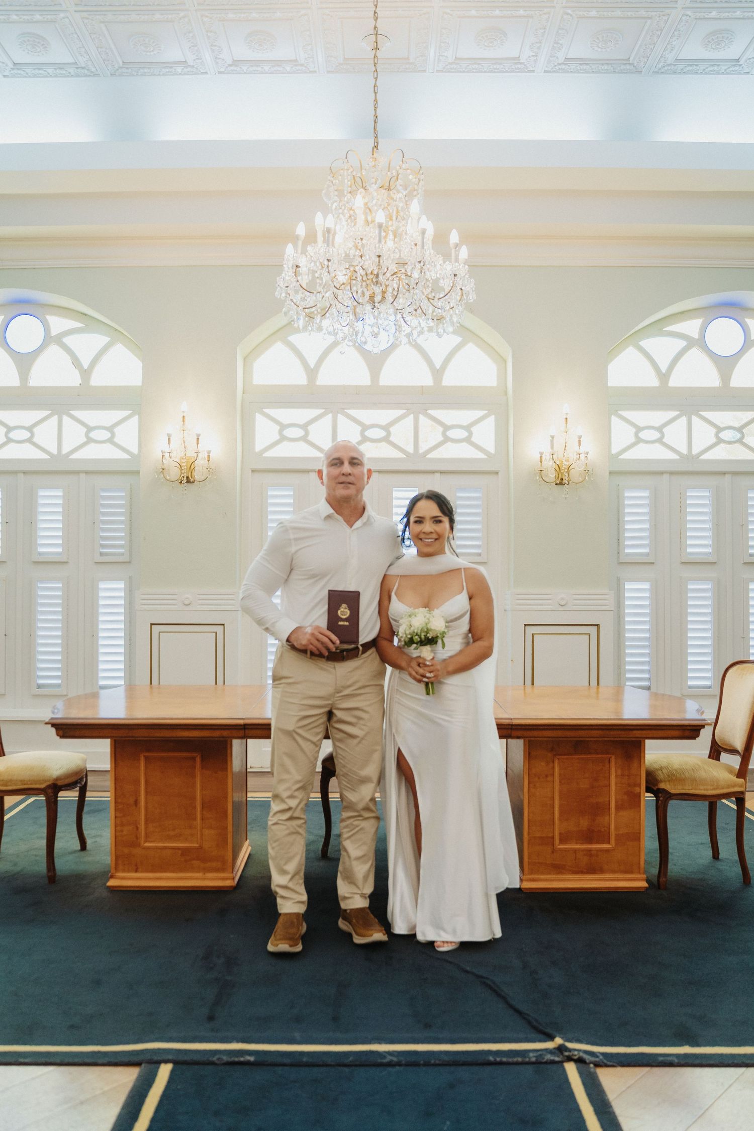 A couple in white wedding attire shares playful moments on a bench in an elegant room with chandeliers and arched windows.