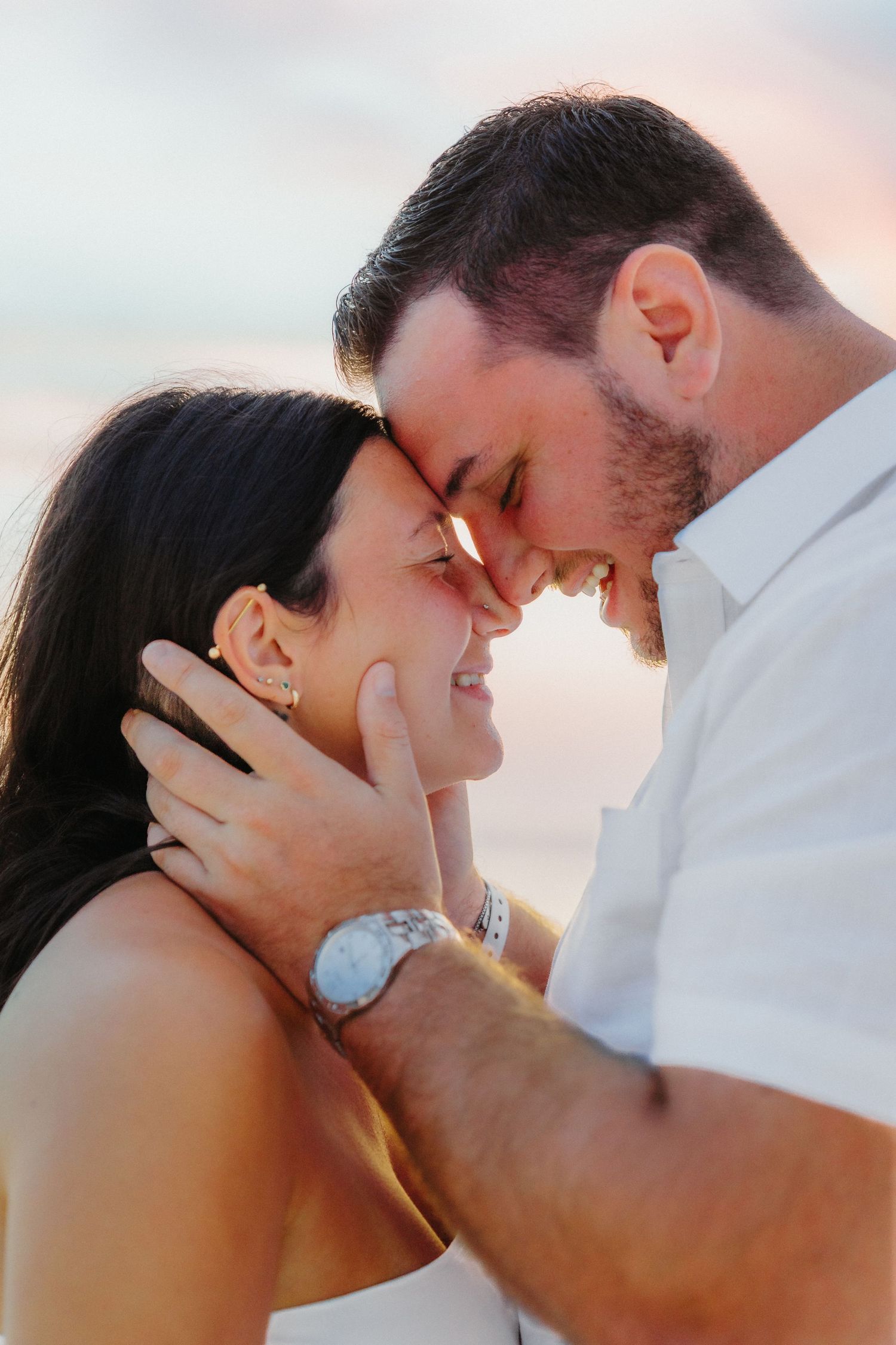 Romantic couple sharing intimate kisses during sunset with soft pink sky in background.
