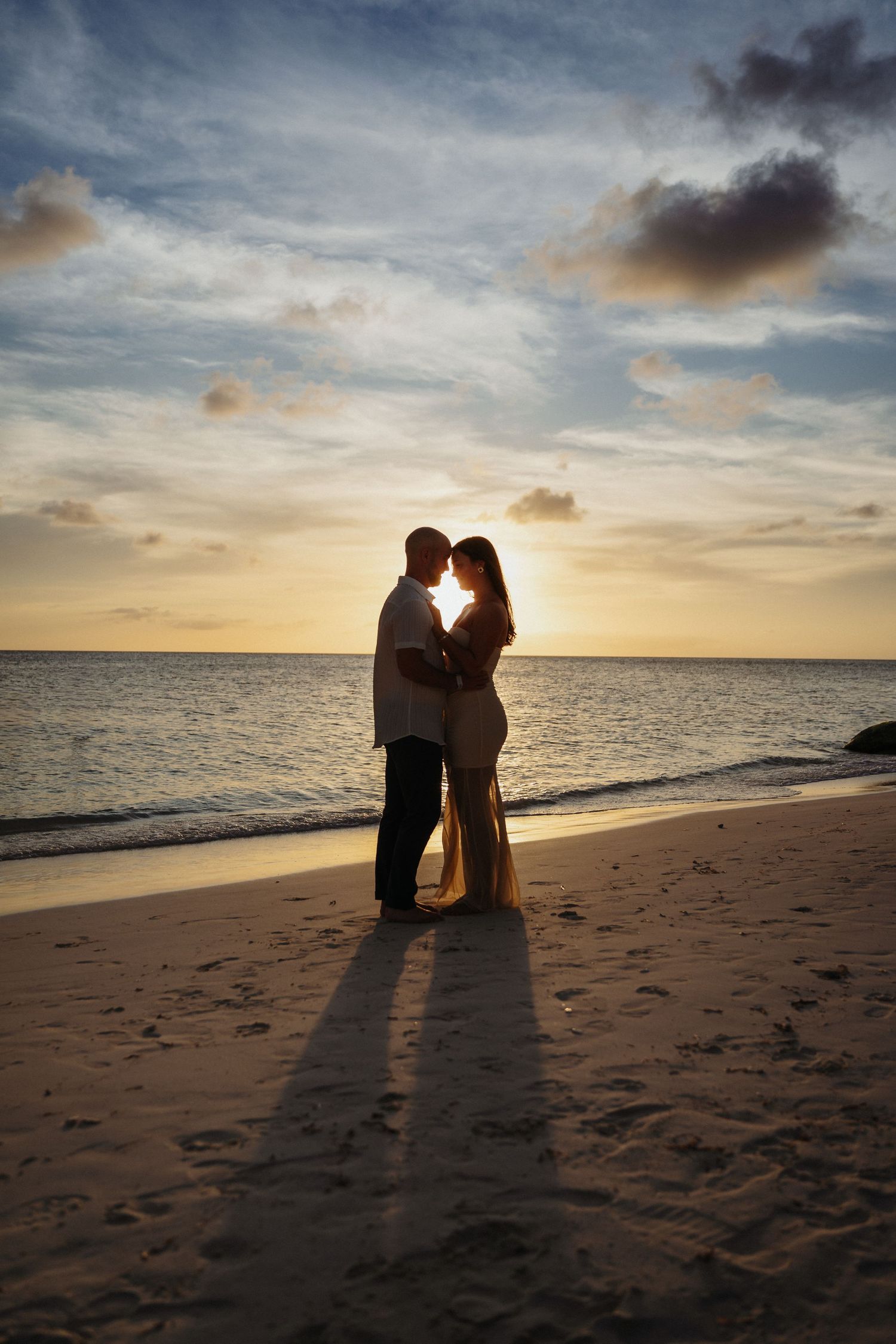 Silhouetted couple sits together on a sandy beach at sunset with dramatic clouds and ocean waves in the background.