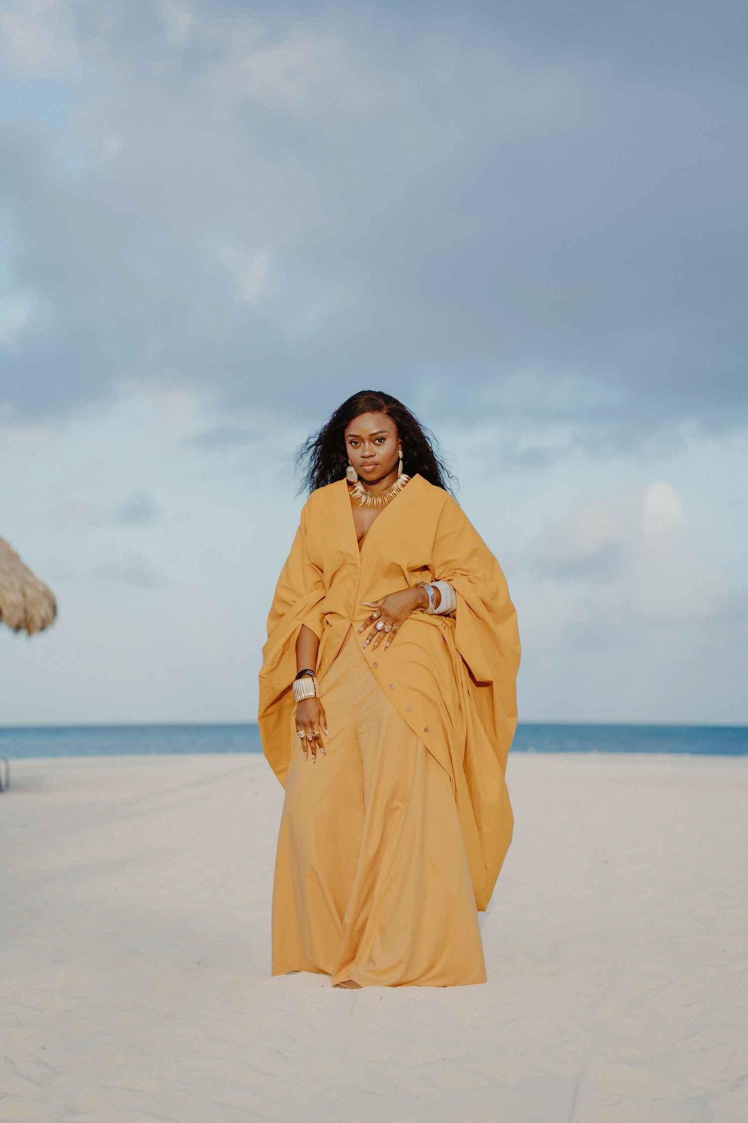 A person in a flowing marigold yellow dress poses gracefully on a pristine white sand beach against a cloudy sky.