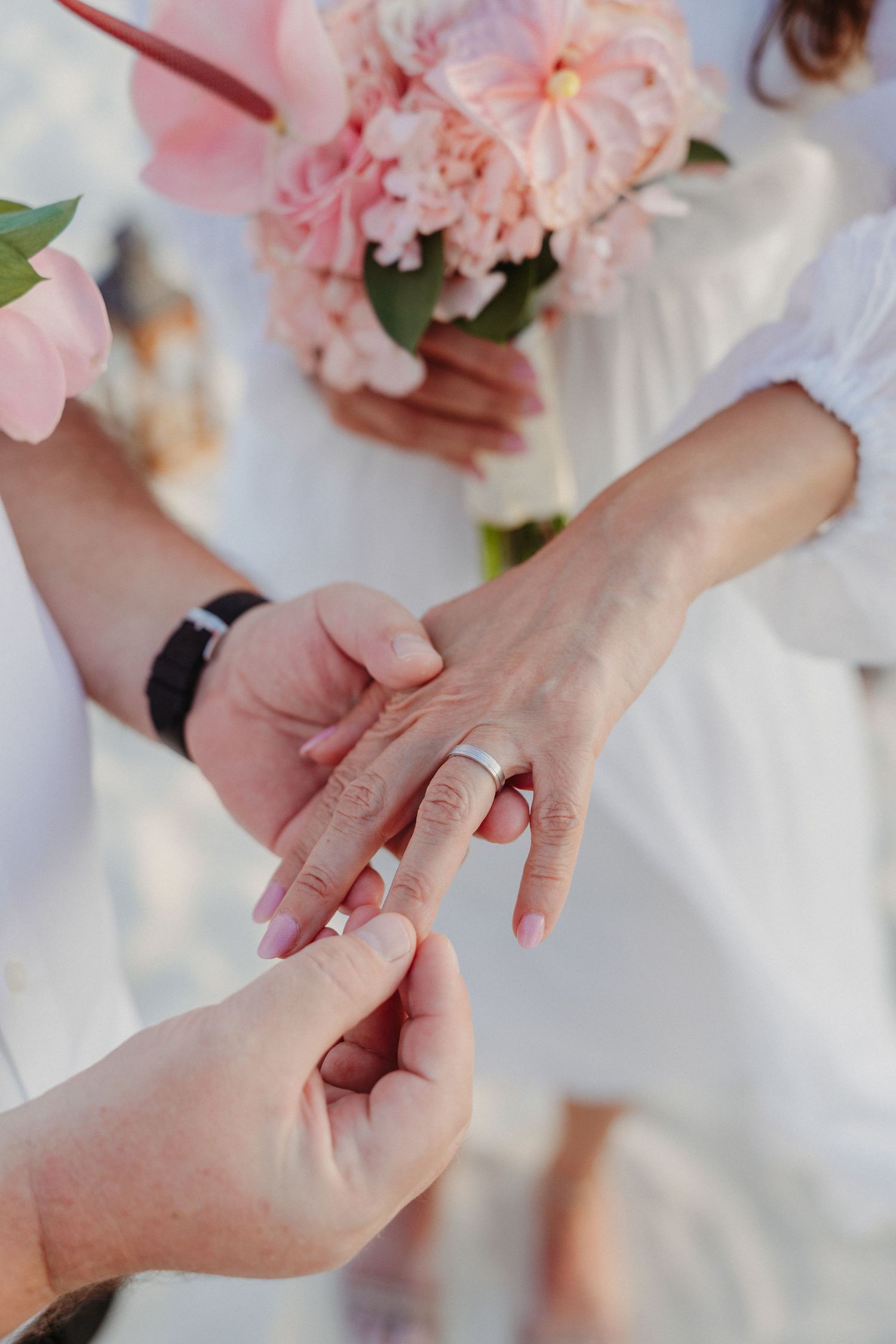 Close-up of hands exchanging wedding rings with soft pink flowers in the background.