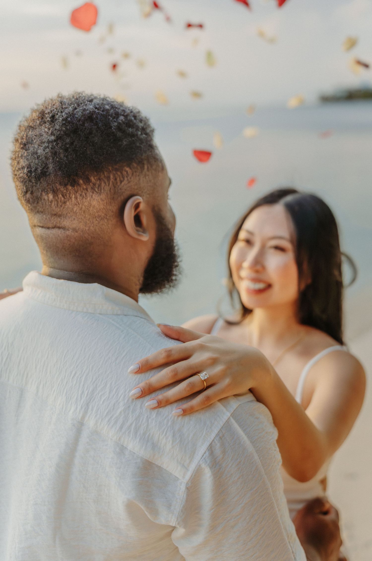 A romantic couple embraces on the beach at sunset with rose petals falling around them in a dreamy atmosphere.