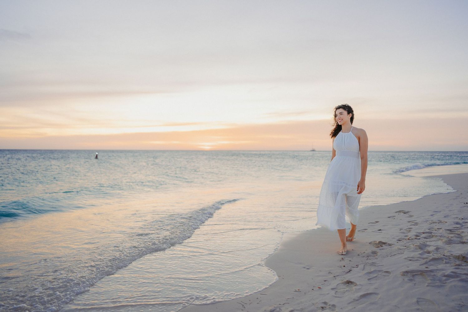 A person in a white dress walks along a serene beach at sunset with gentle waves lapping at the shore.
