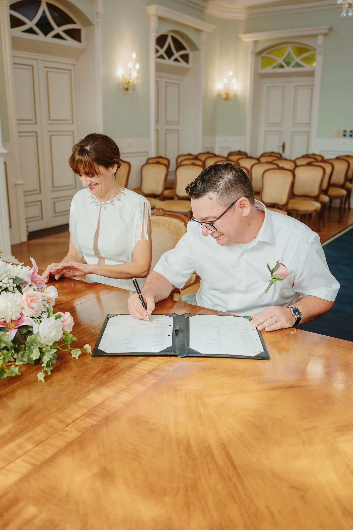 Multiple people in white clothing sign documents at a wooden table in an elegant room with ornate wall sconces.