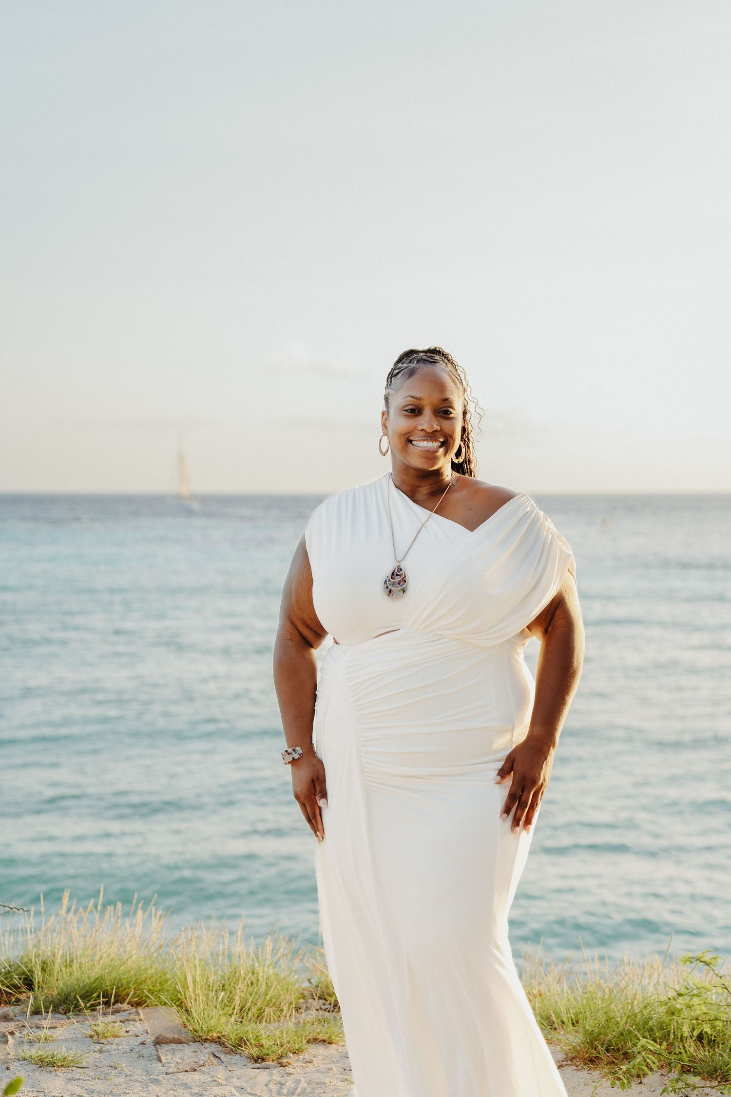 A figure in a flowing white dress poses gracefully against a serene ocean backdrop at sunset.