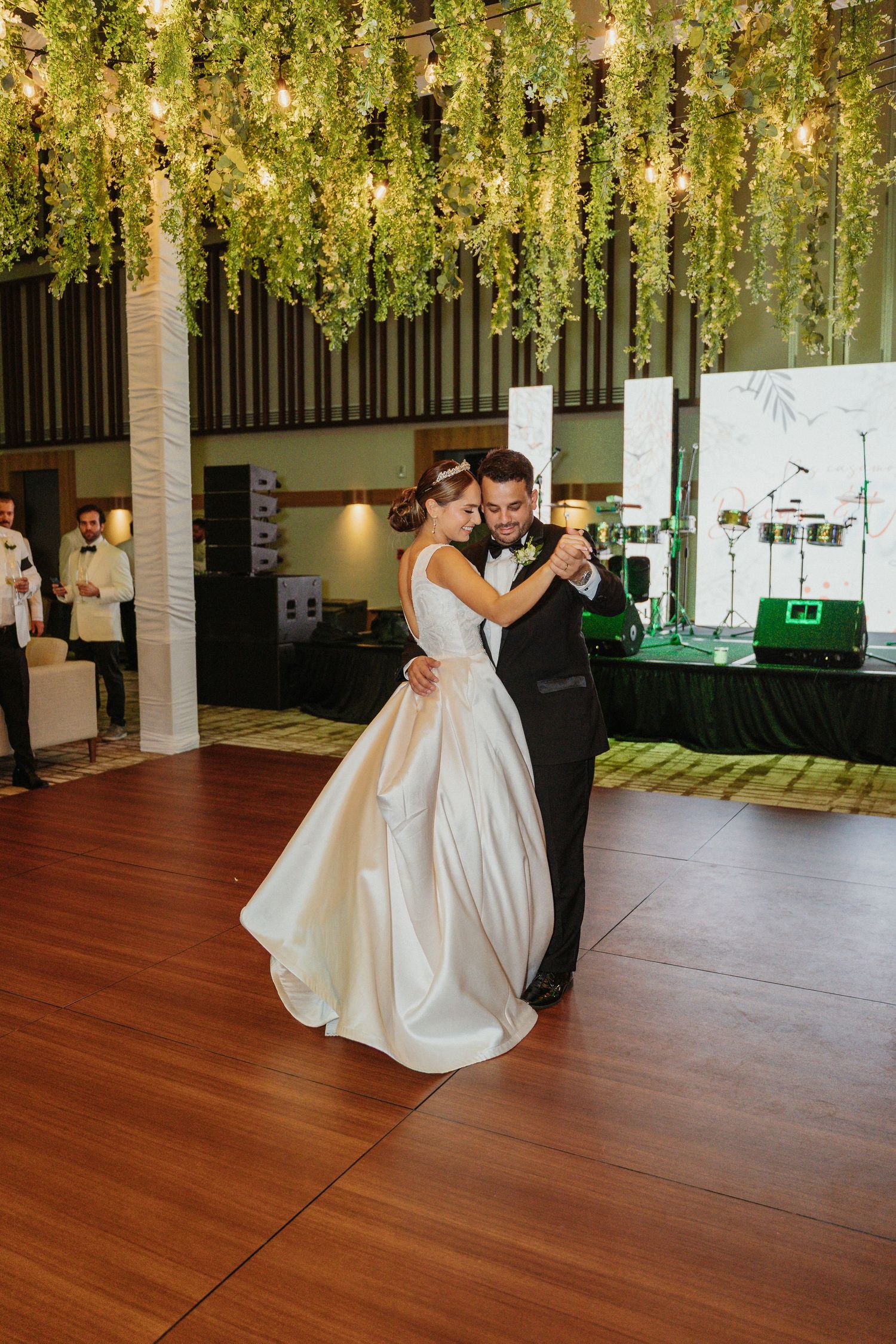 A couple shares a romantic dance on a wooden floor under hanging floral decorations at a wedding reception.