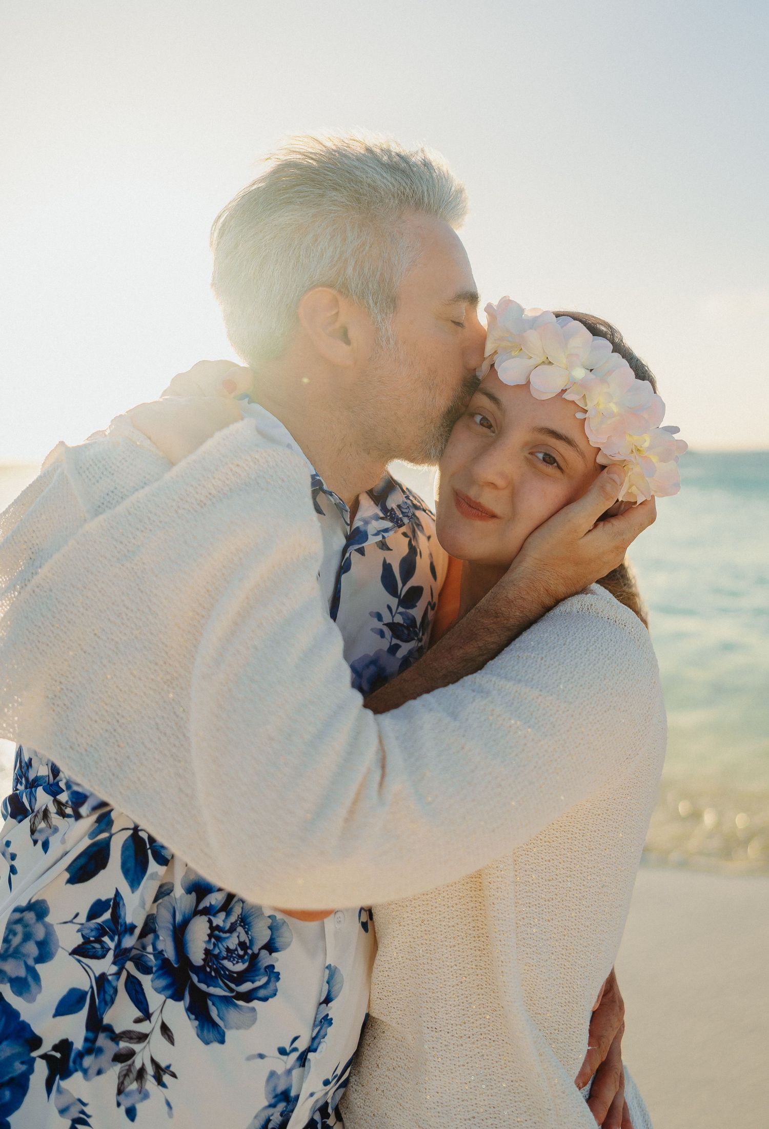 A romantic couple in white outfits embracing on the beach at sunset with soft lighting and floral details.