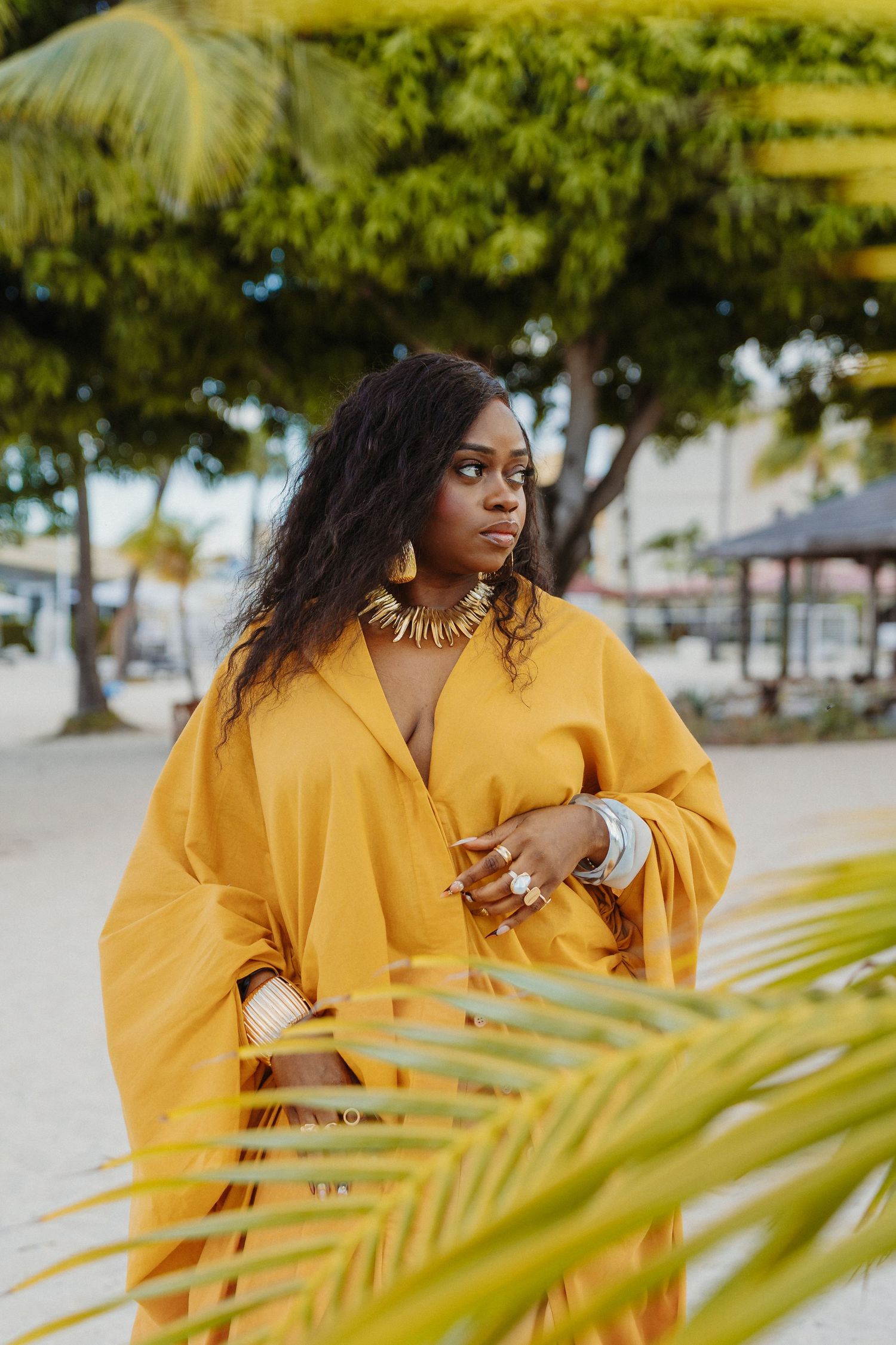 Woman in bright yellow sweater poses near tropical palm fronds at a beach location.
