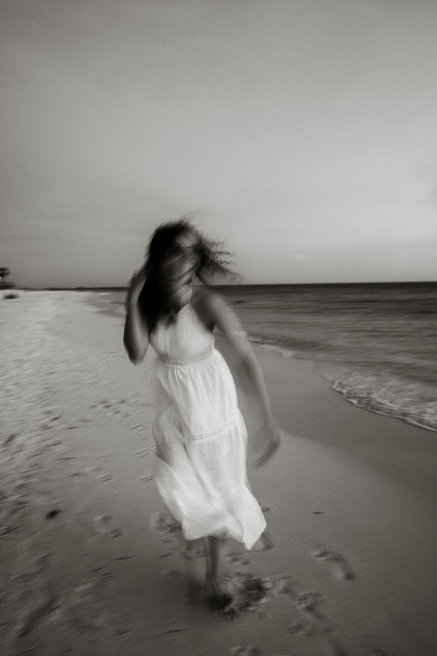 A figure in a flowing white dress twirls on a beach at dusk, creating a dreamy black and white scene.