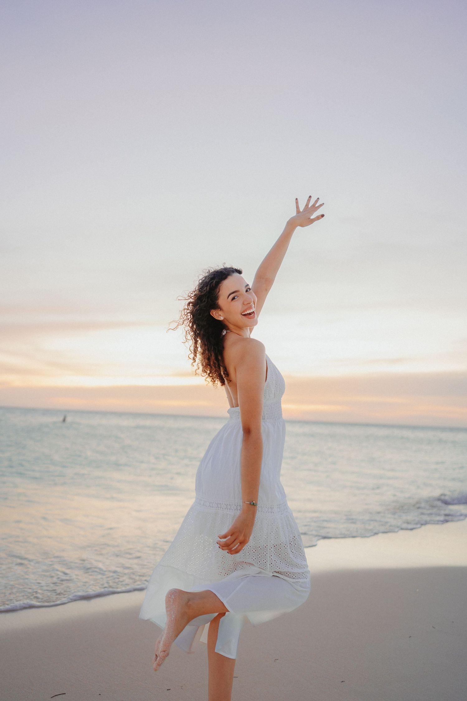 A person in a flowing white dress dances joyfully along a serene beach at sunset.