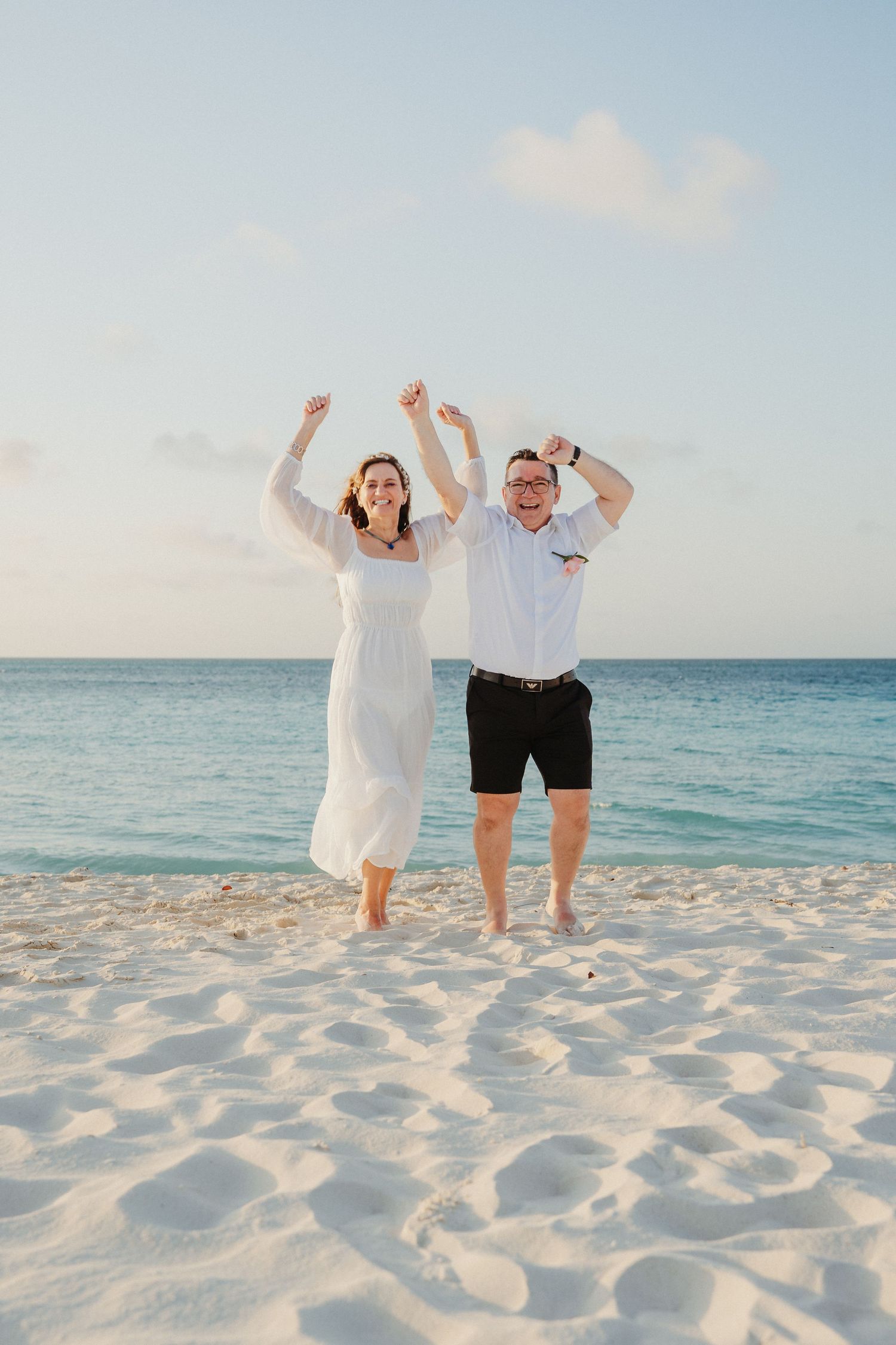 A couple celebrates joyfully on a pristine white sand beach with turquoise ocean waters in the background.