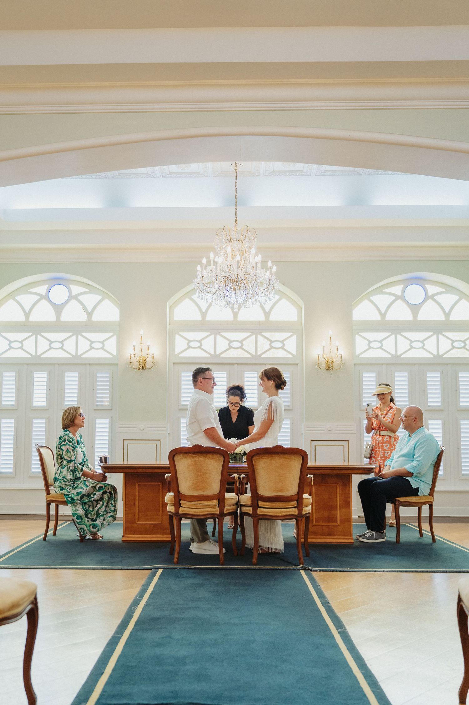Small intimate wedding ceremony in an elegant room with arched windows, crystal chandelier and teal carpet aisle.
