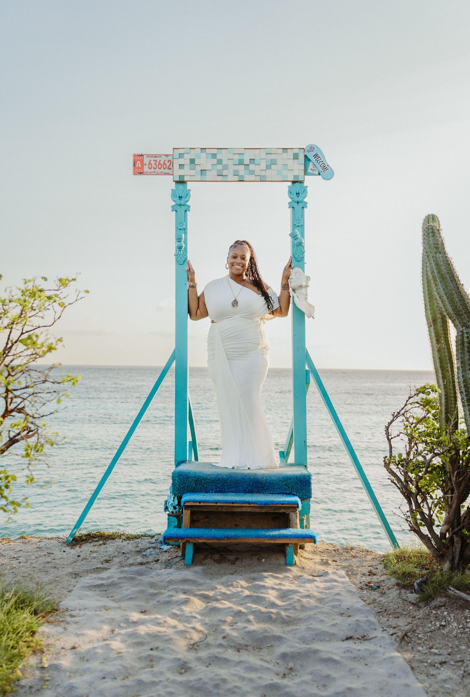 A series of turquoise wooden beach swings overlooking the ocean with cacti and coastal vegetation on a sandy shore.