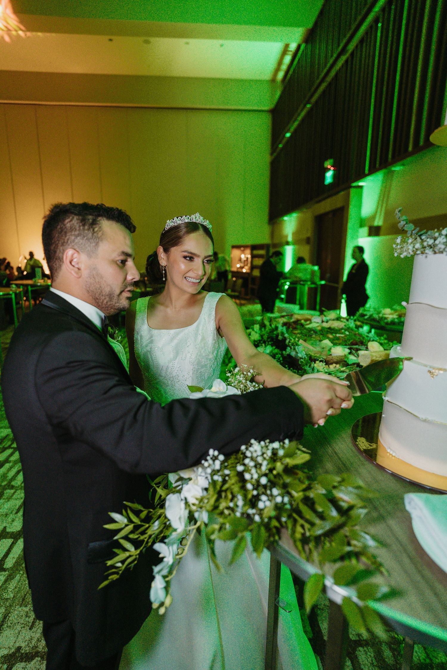 A couple cuts into their elegant white wedding cake decorated with greenery under green ambient lighting.