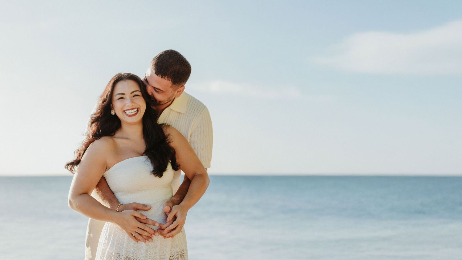 A couple embraces lovingly on a sunny beach with crystal clear blue ocean waters in the background.