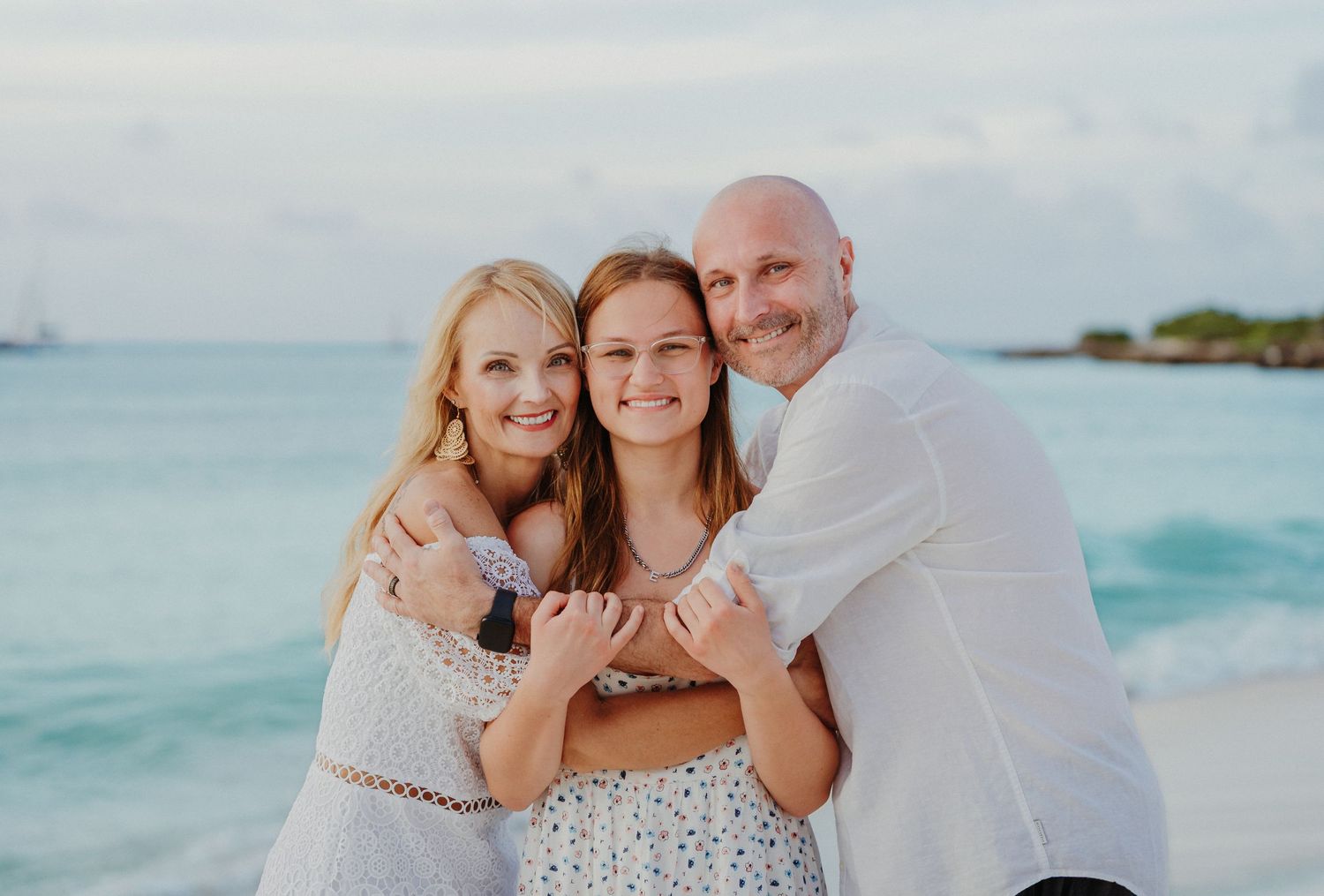 Three people pose together on a tropical beach with turquoise water and palm trees in the background.