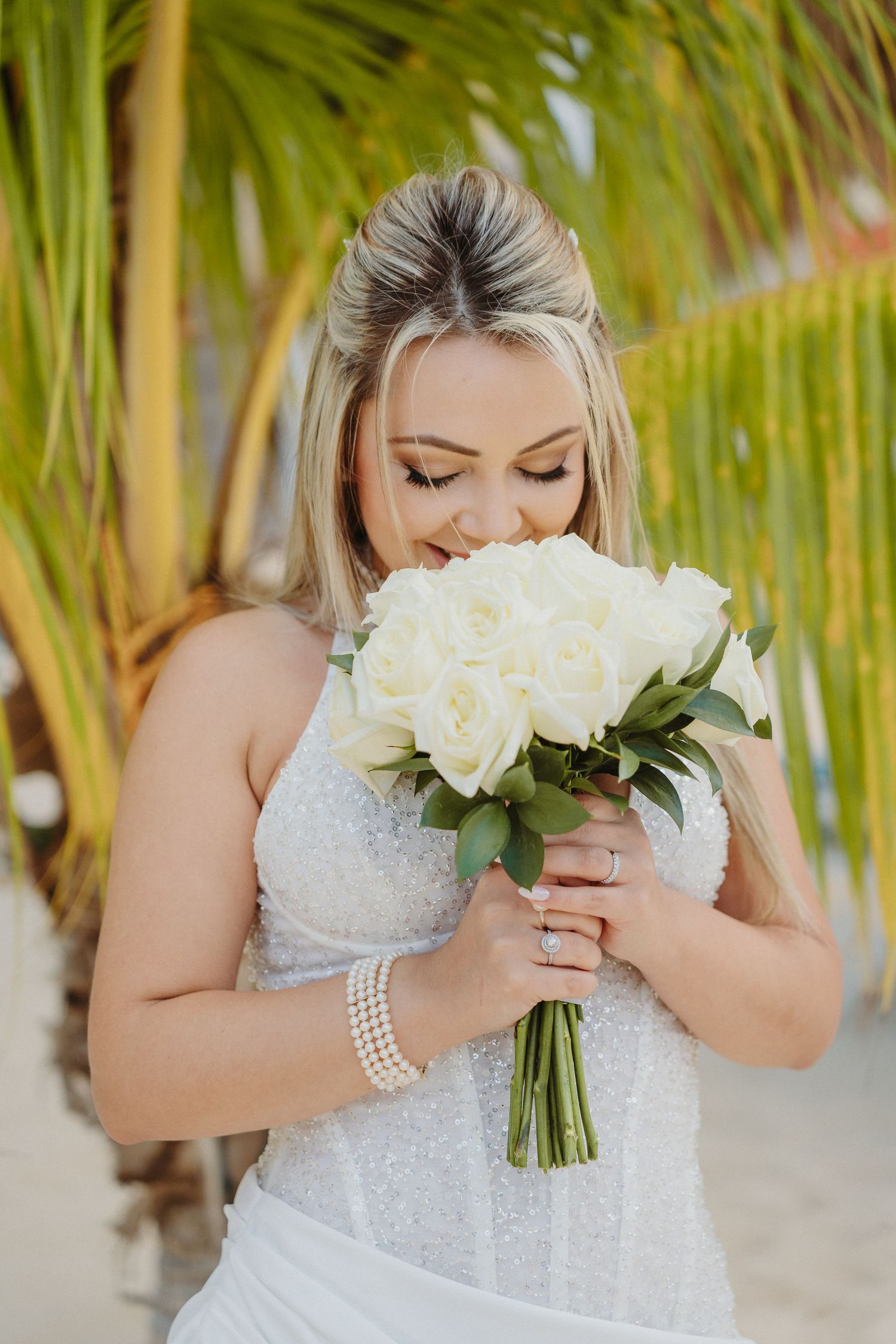 Bride in white lace dress holds elegant white rose bouquet against tropical palm background at beach wedding.