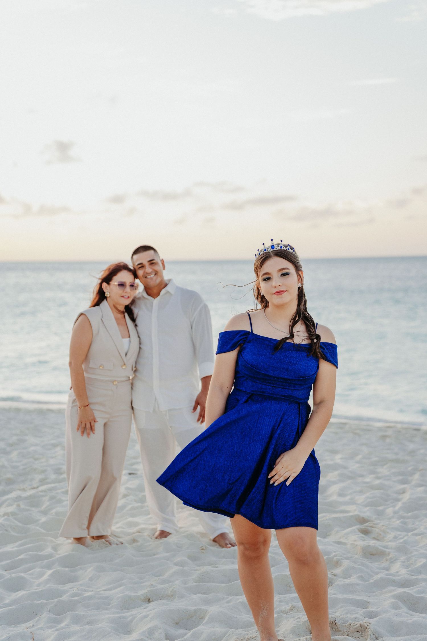 Group poses playfully on a white sandy beach at sunset wearing blue and beige formal attire.