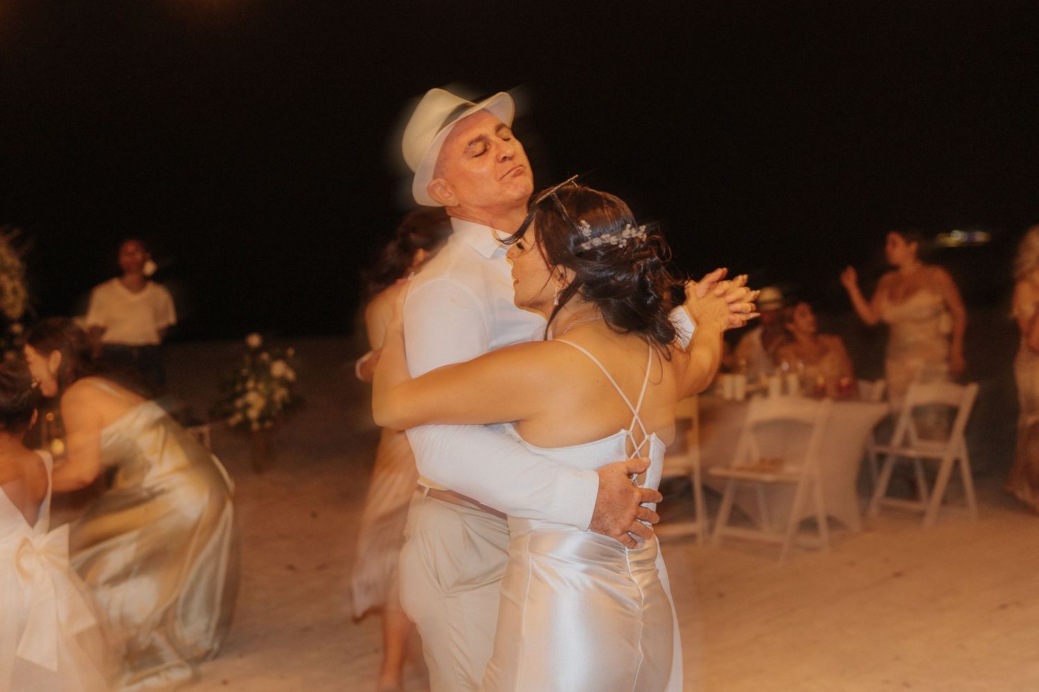 A couple dances together at an outdoor evening wedding reception with white chairs and guests in the background.