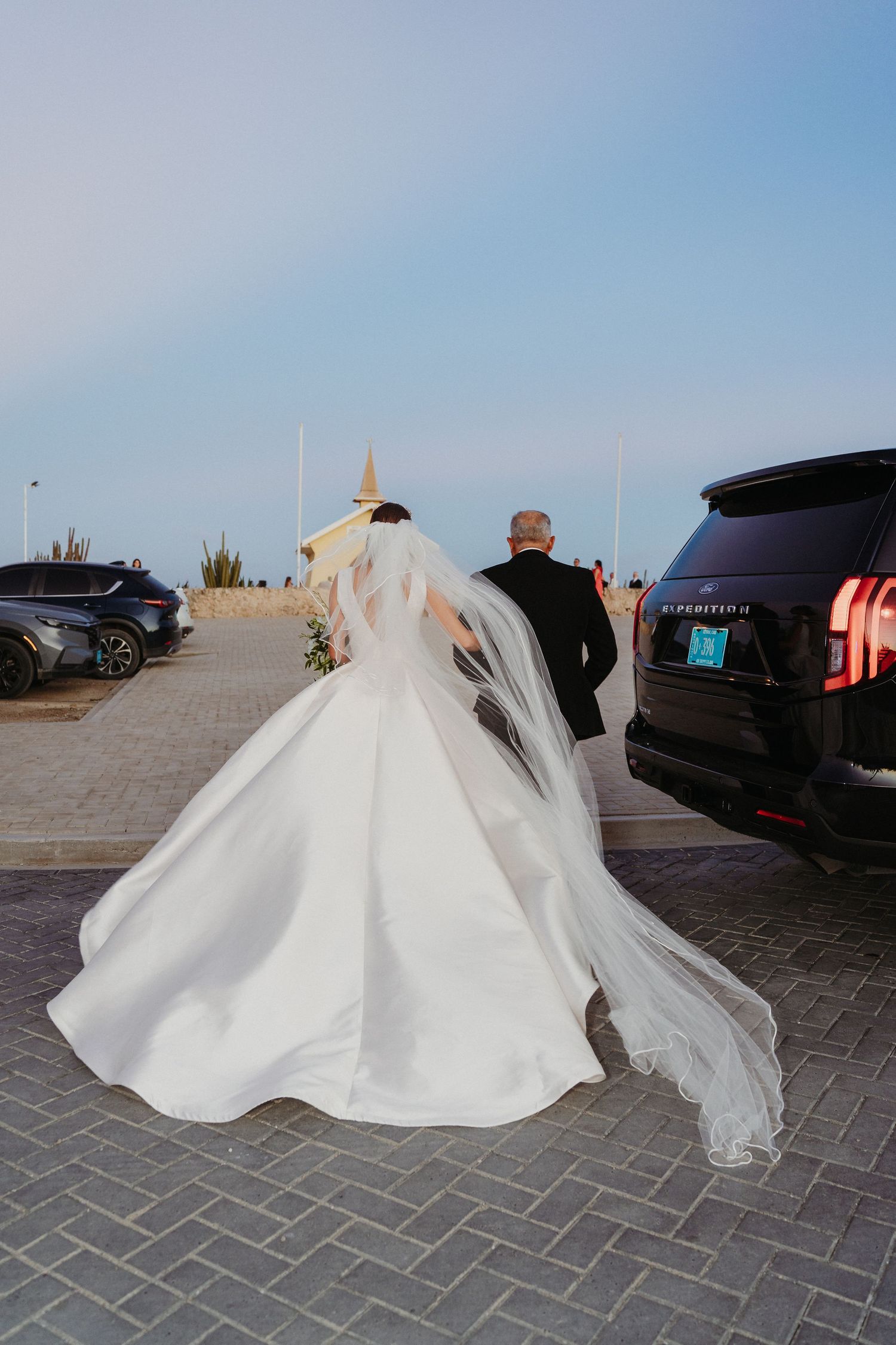 A bride in a flowing white wedding gown with a long veil walks across a brick paved area near parked vehicles.