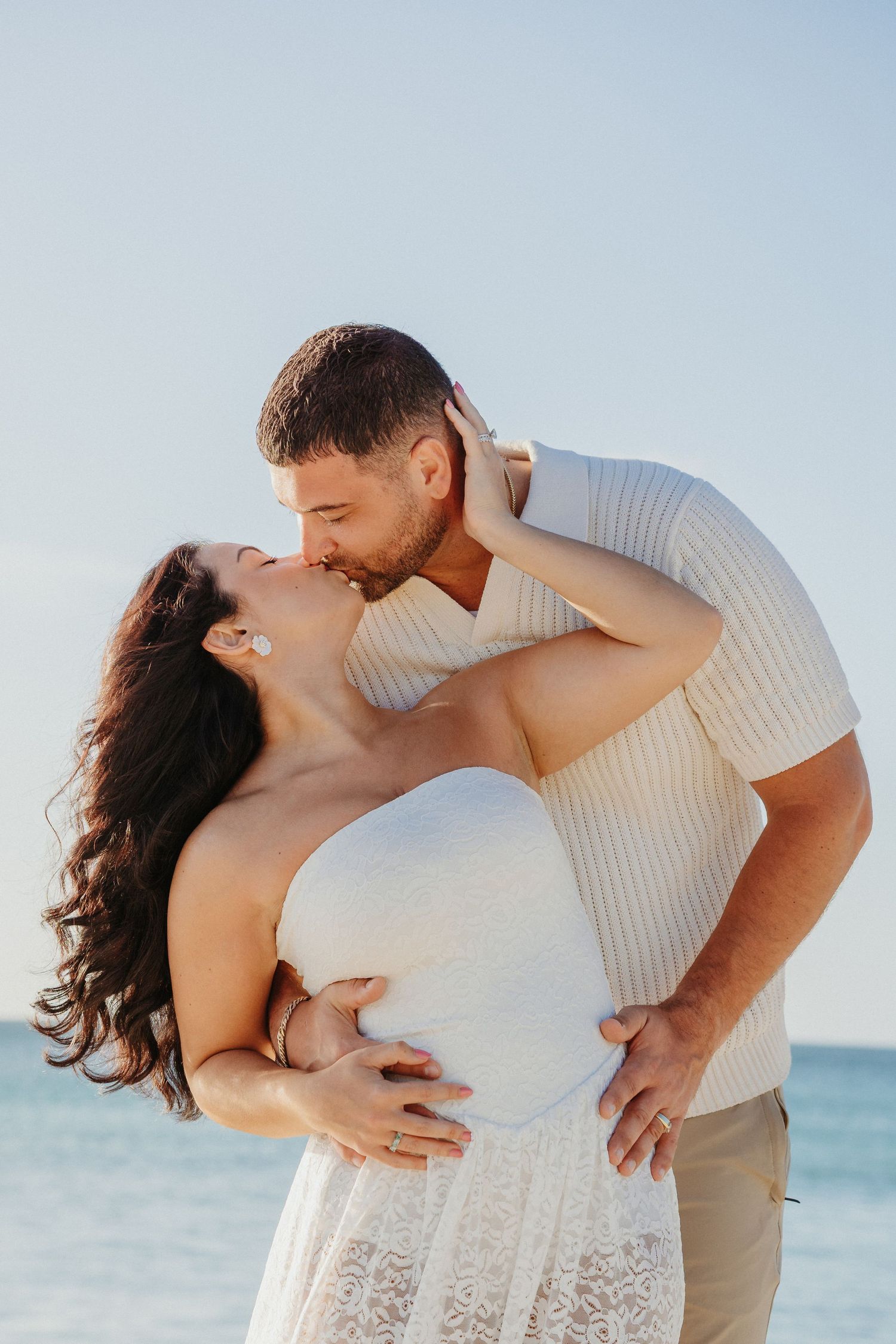 A romantic couple shares tender moments on a sunny beach while wearing light colored outfits.