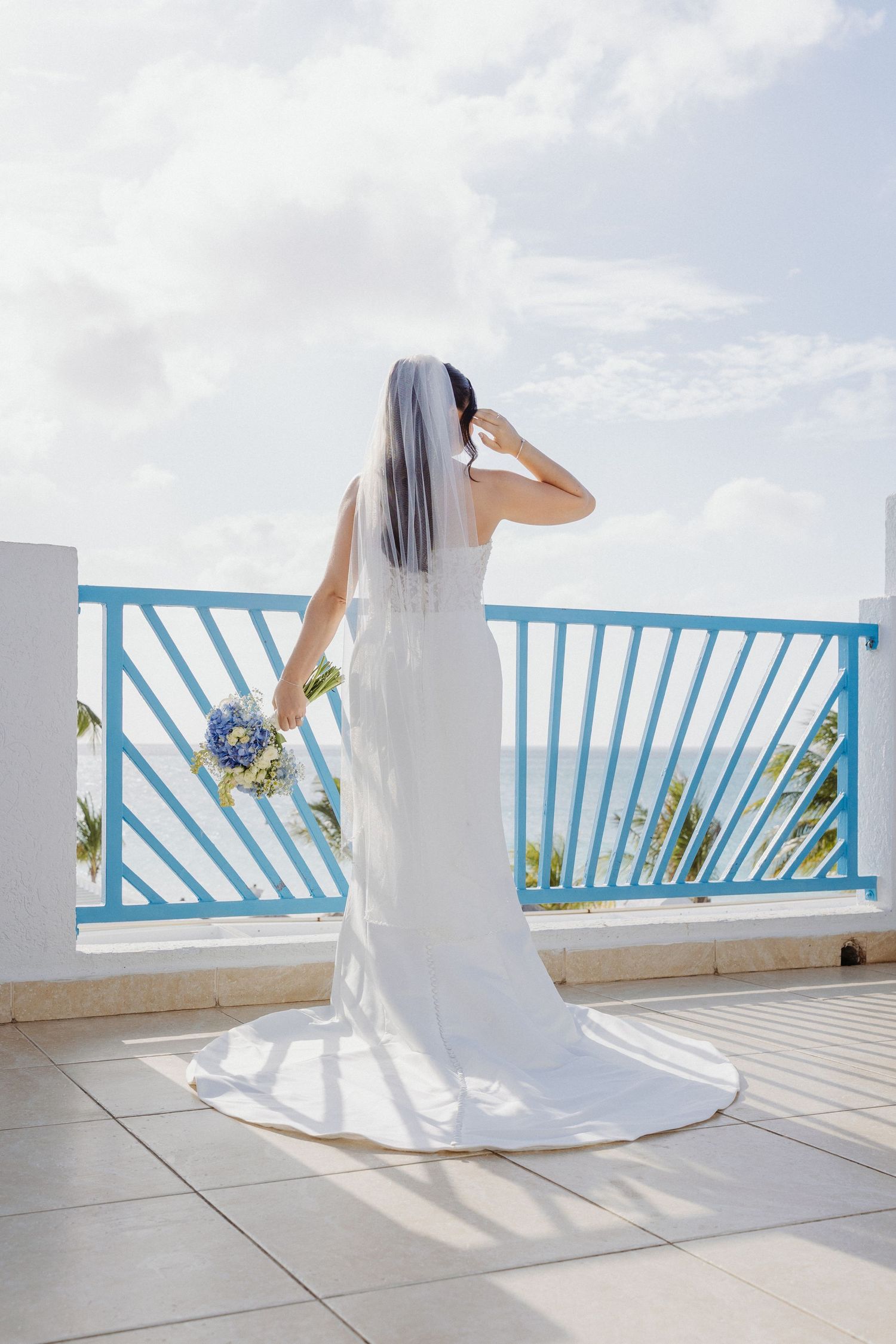 Bride in white gown and veil holds blue bouquet on ocean-view terrace with turquoise railing.