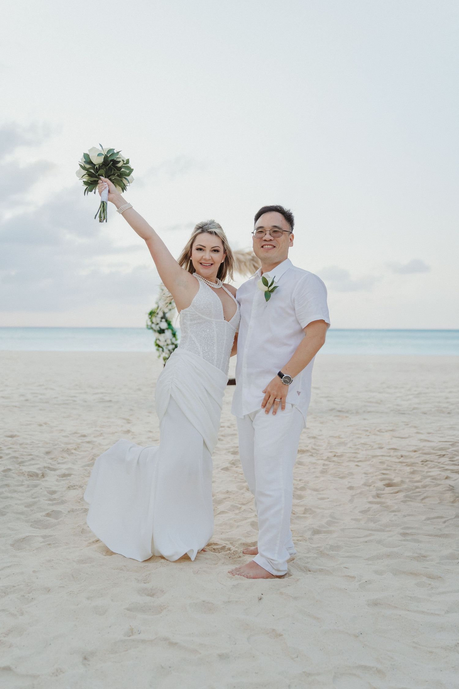 Joyful wedding couple poses playfully on tropical beach with white attire and ocean backdrop.