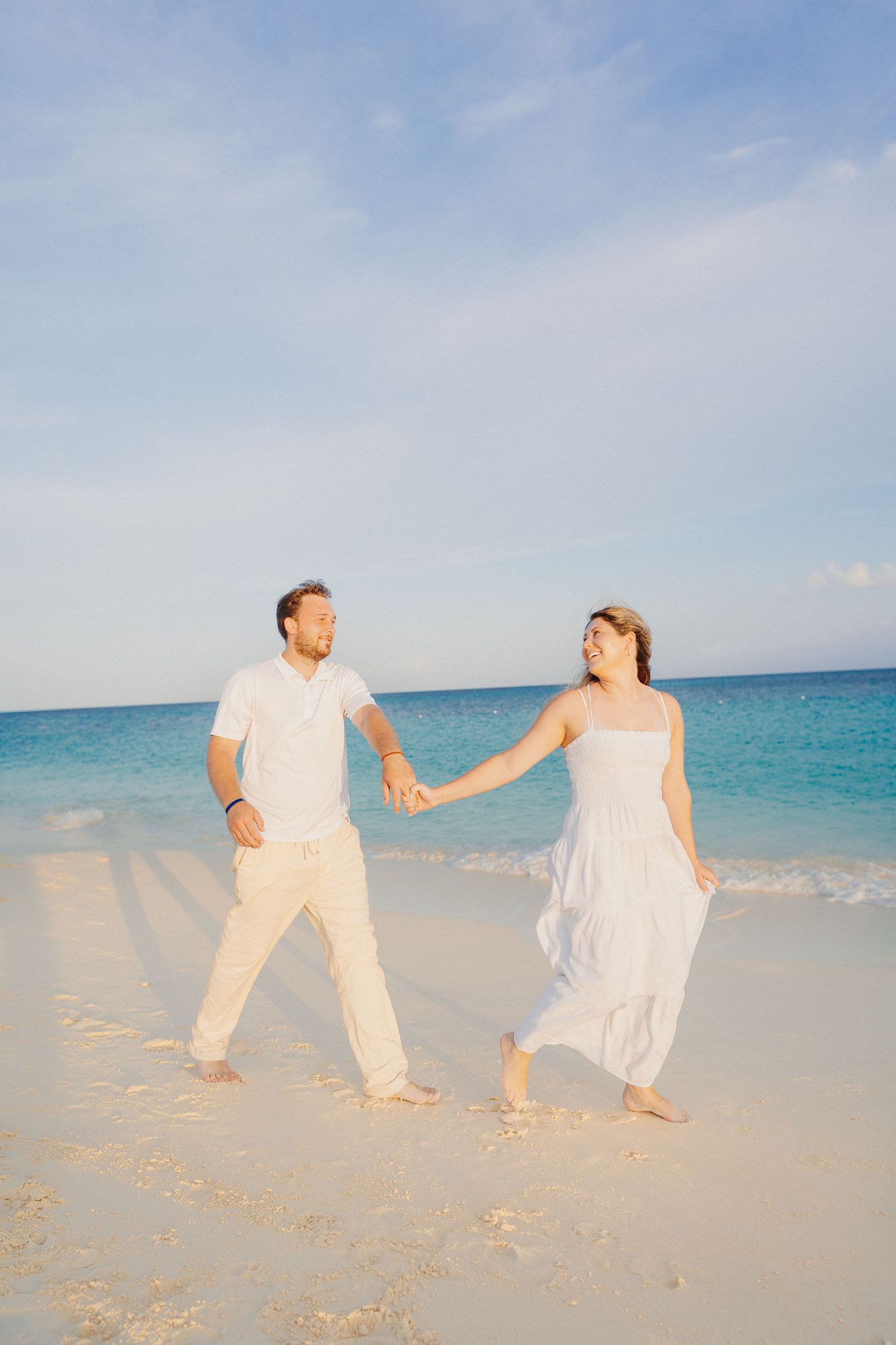 A couple in white and peach attire dances playfully on a tropical beach with turquoise ocean waters in the background.