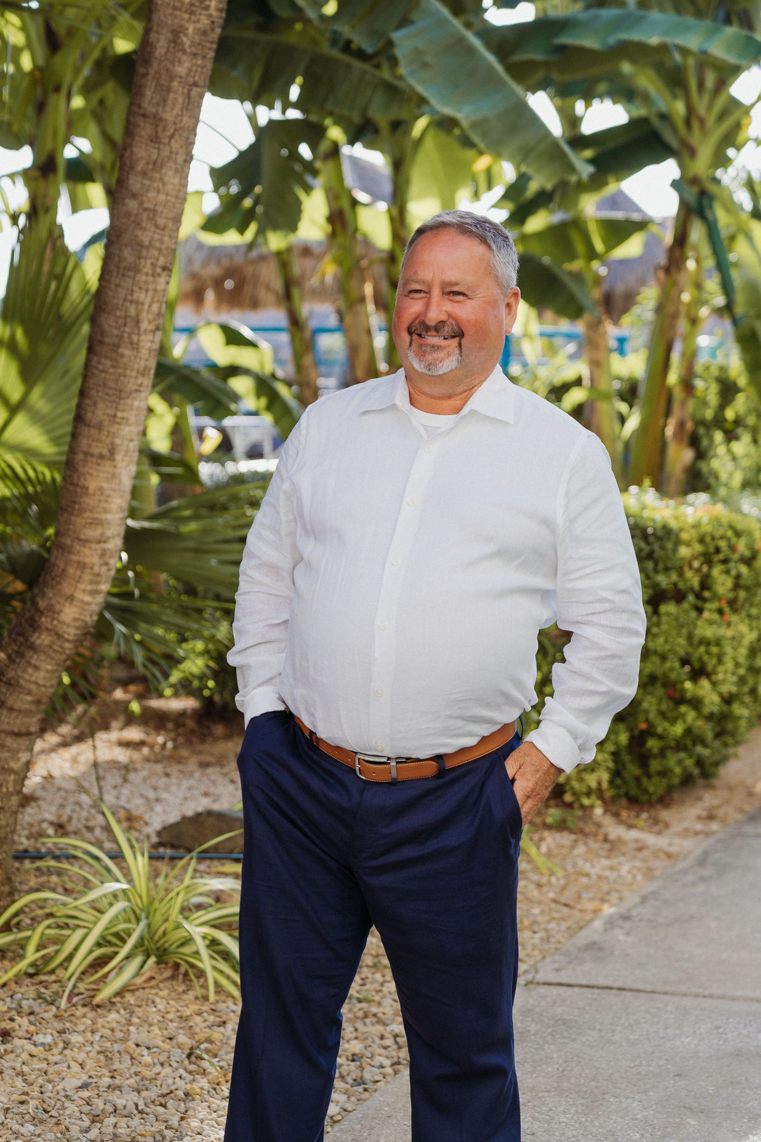 Smiling middle-aged man in white shirt and navy pants standing outdoors among tropical palm trees and lush greenery.
