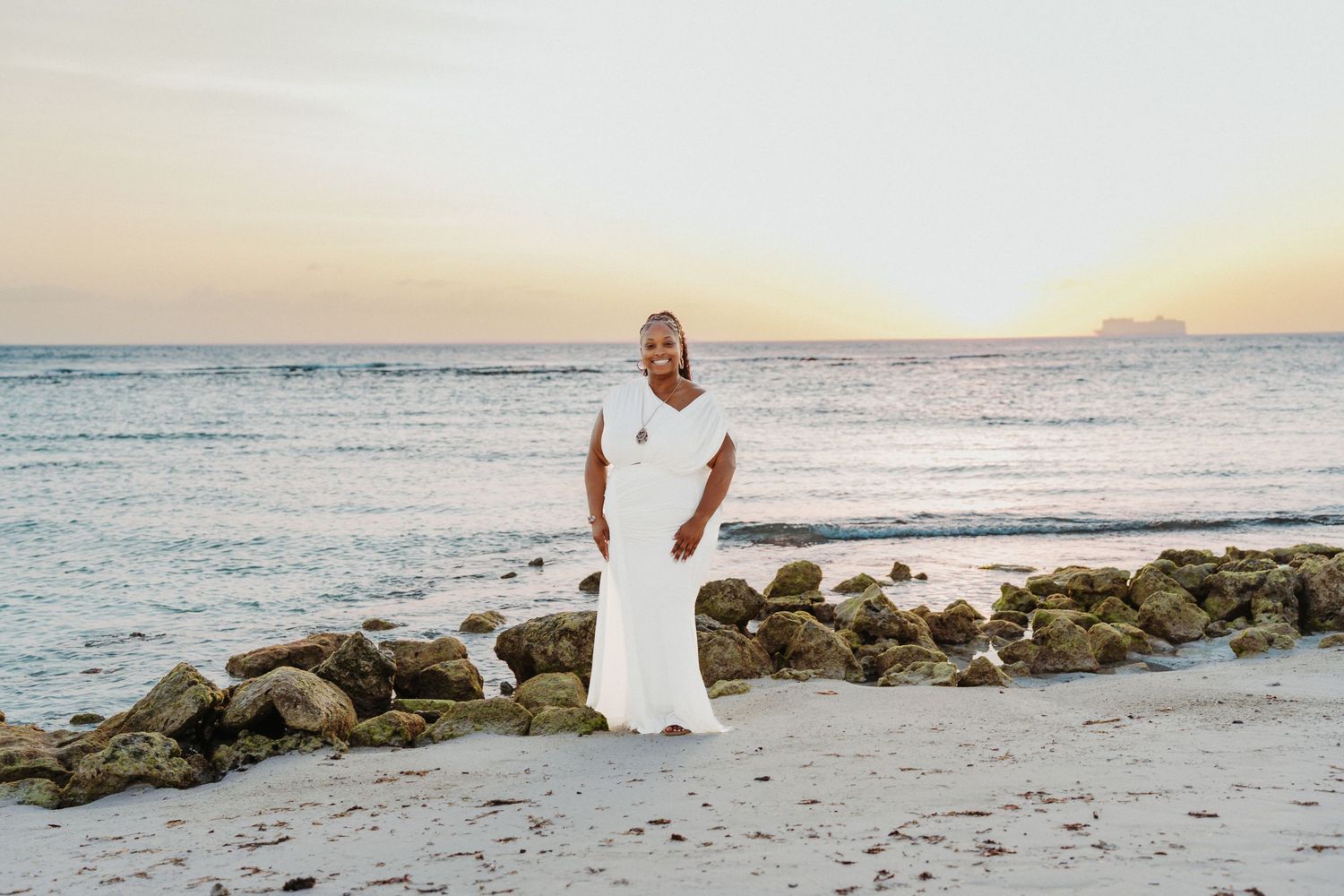 Figure in white dress stands on beach at sunset with rocky coastline and ocean waves in background.