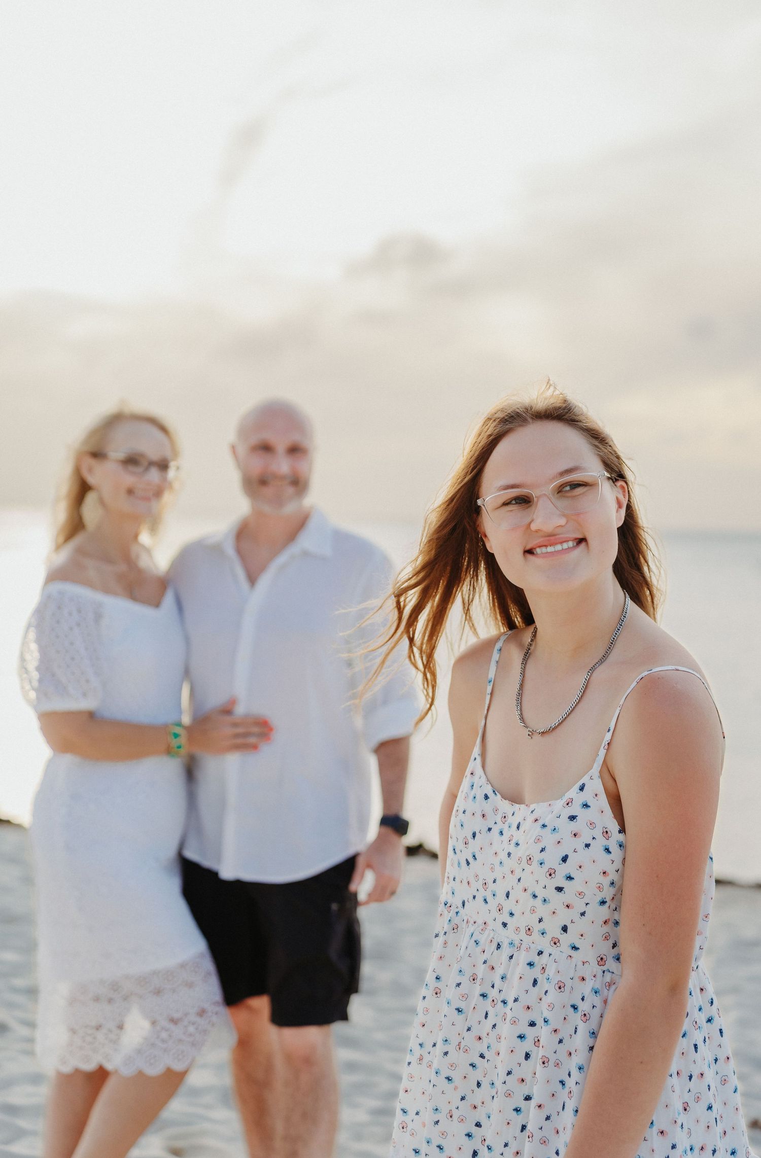 A family walks and poses together on a sunny beach wearing white and floral summer outfits during a photo session.