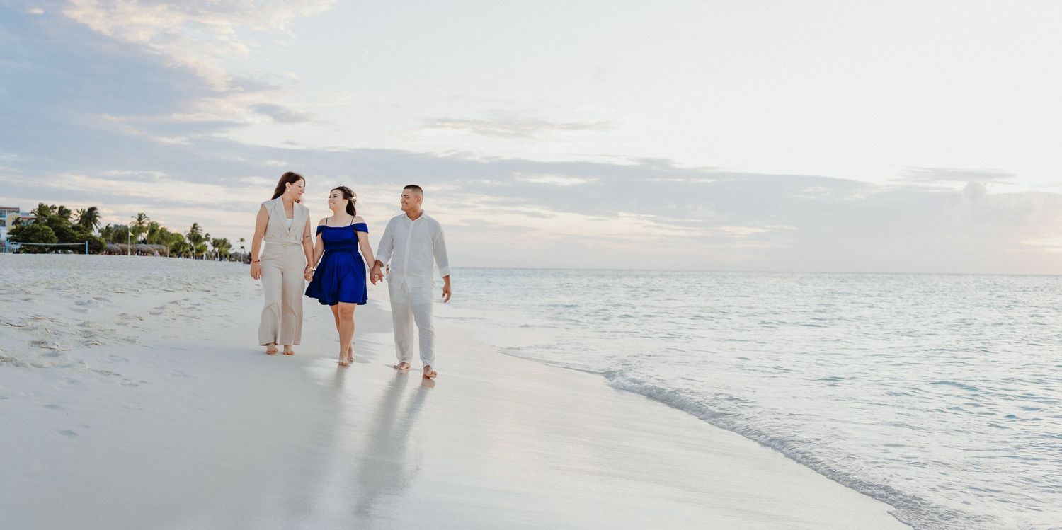 A couple walks along a pristine white sand beach at sunset with palm trees and ocean waves in the background.