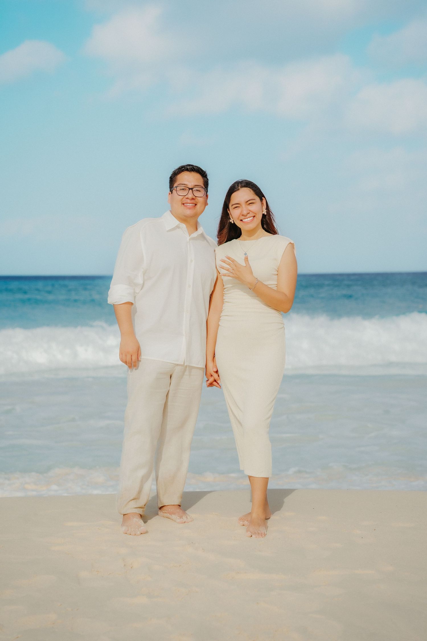 A couple in matching white outfits stands barefoot on a tropical beach with turquoise ocean waves and blue sky behind them.