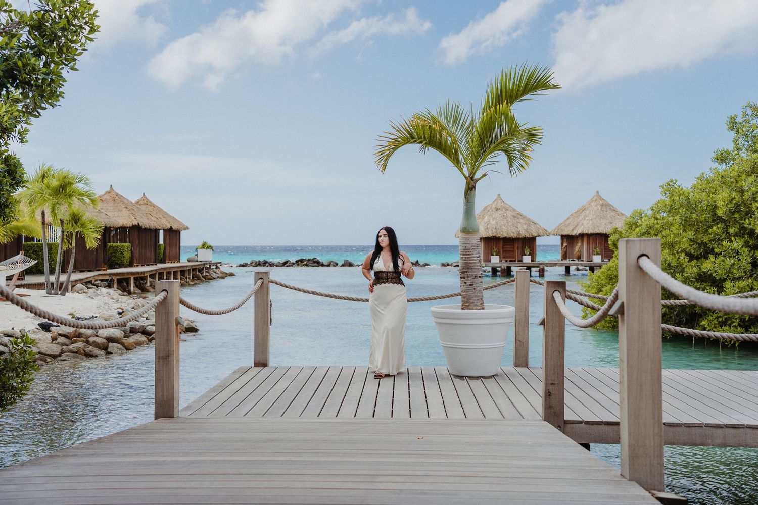 A wooden boardwalk leads to overwater bungalows at a tropical resort with palm trees and turquoise ocean views.