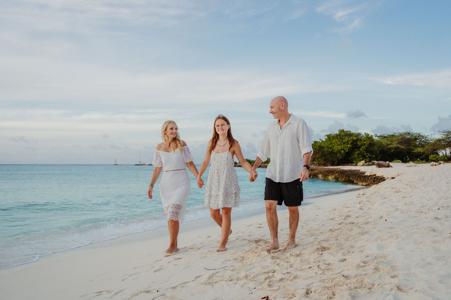 Three people in white outfits walk hand in hand along a tropical beach with turquoise water and blue sky.