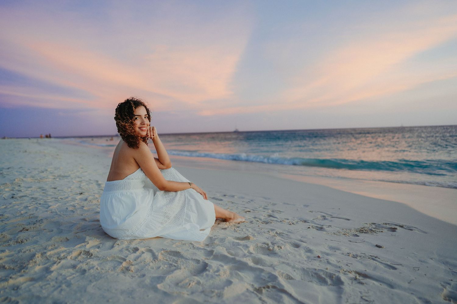 Person in white dress sits on pristine beach at sunset with turquoise ocean waters and pink sky in background.