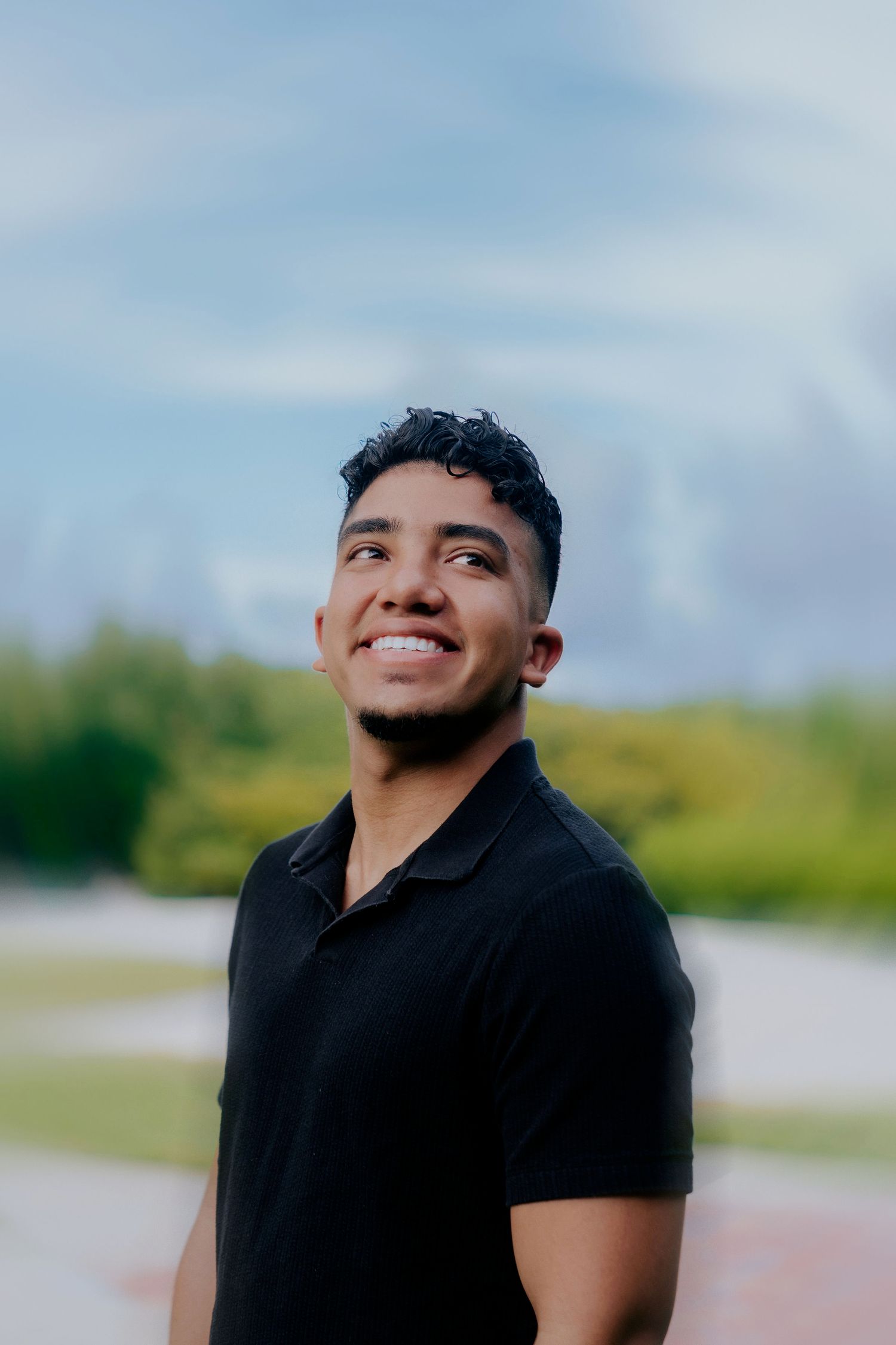 Person in black t-shirt smiling outdoors against natural background with cloudy sky.