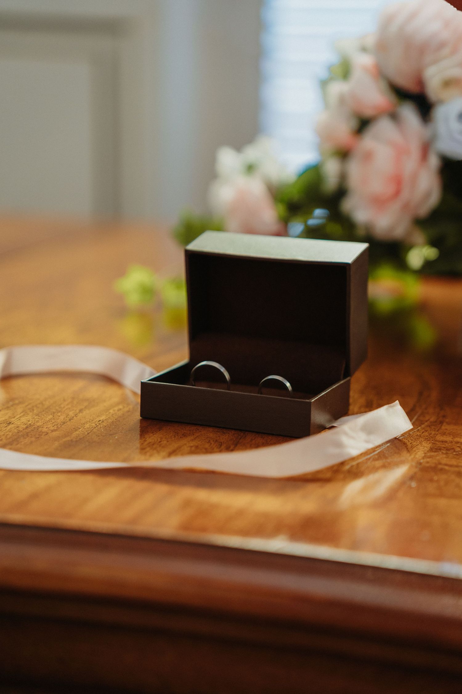 Black jewelry box sits on wooden table with pink roses in background, creating elegant wedding detail shot.