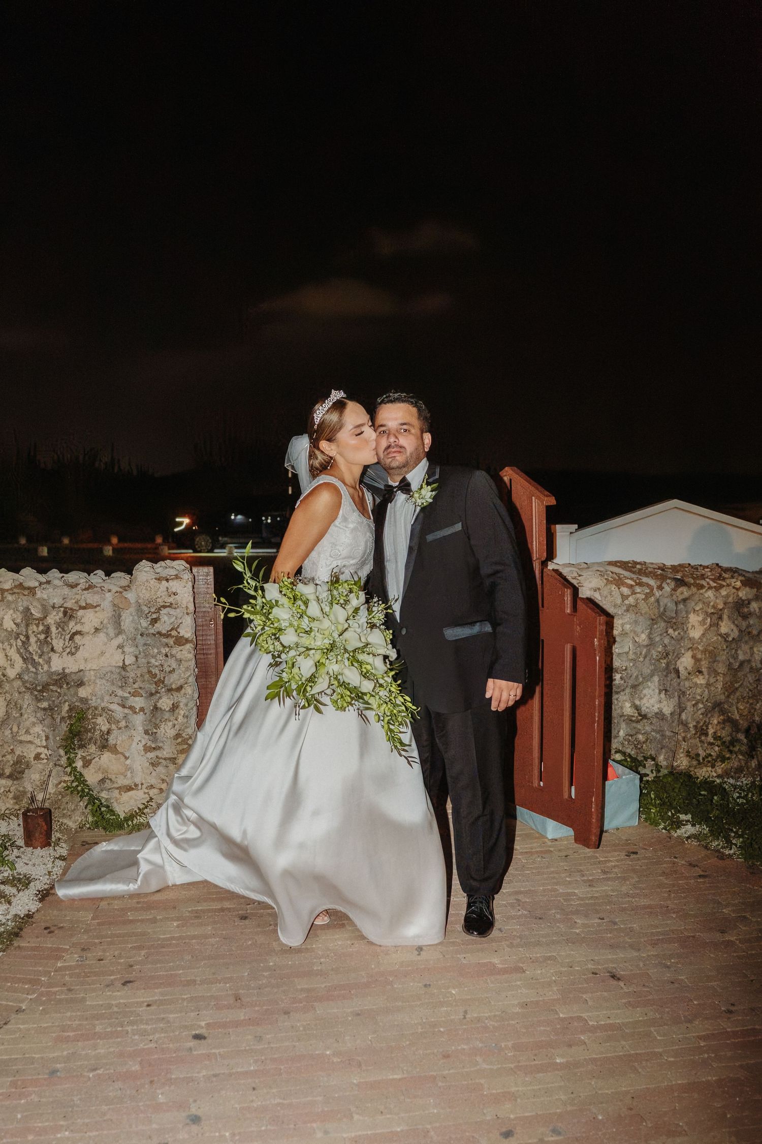 A wedding couple shares a romantic moment at night against a stone wall backdrop with a bouquet of white flowers.