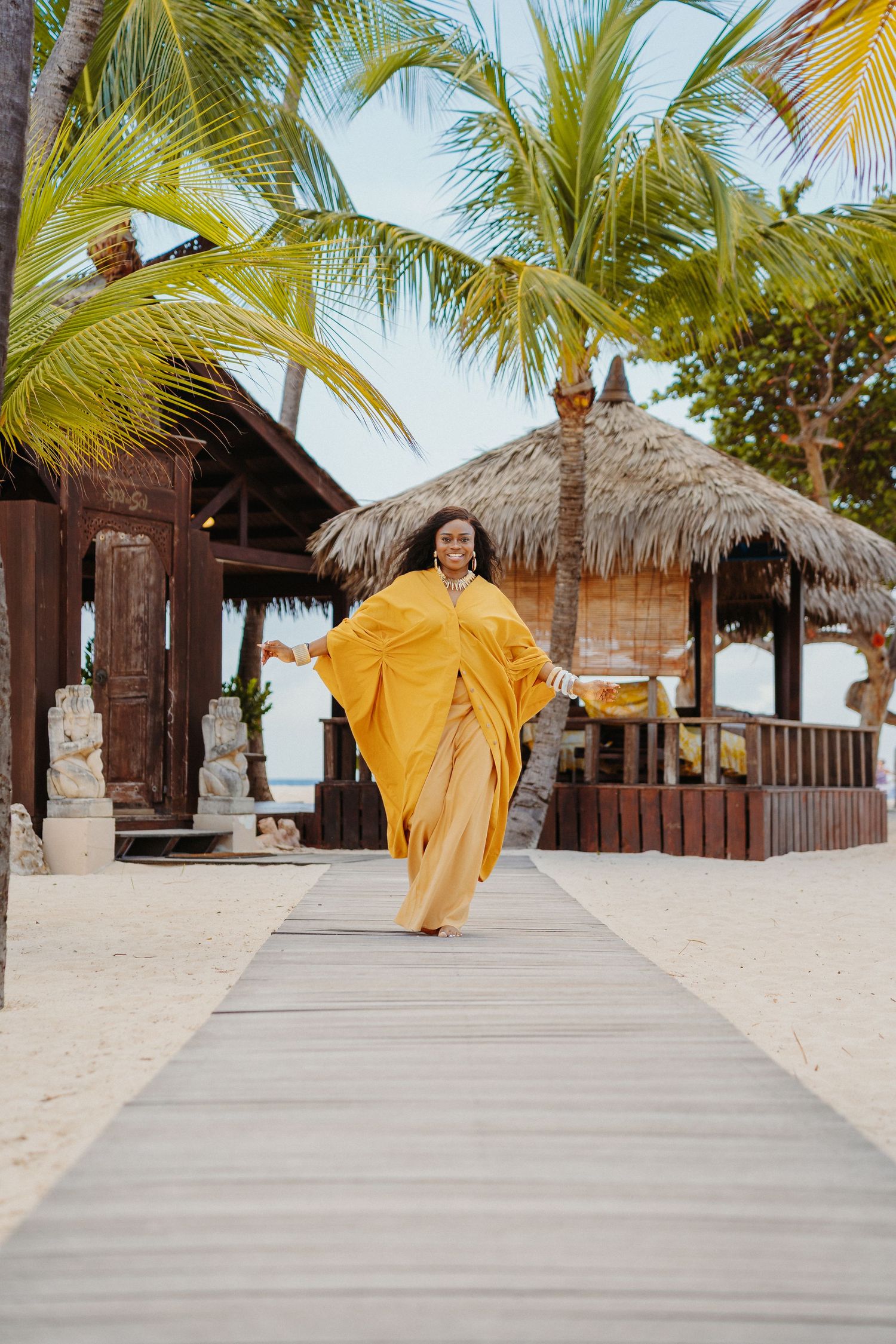 Person in flowing yellow dress twirls joyfully on a tropical beach with palm trees and thatched huts in the background.