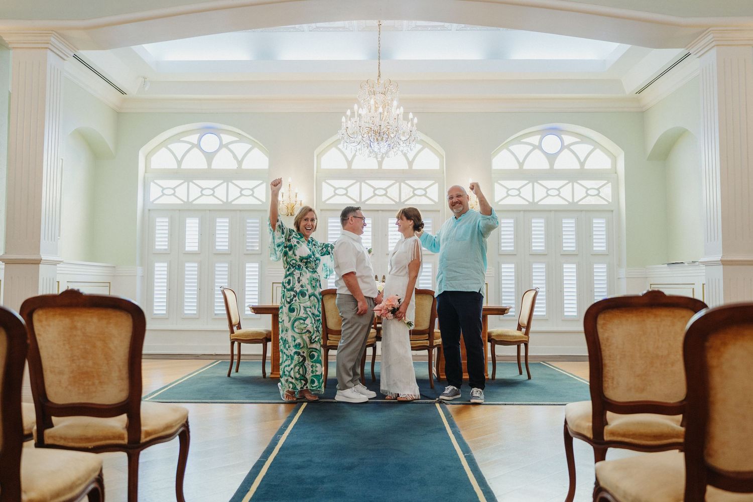 A group celebrates in an elegant room with arched windows, a chandelier, and antique chairs.