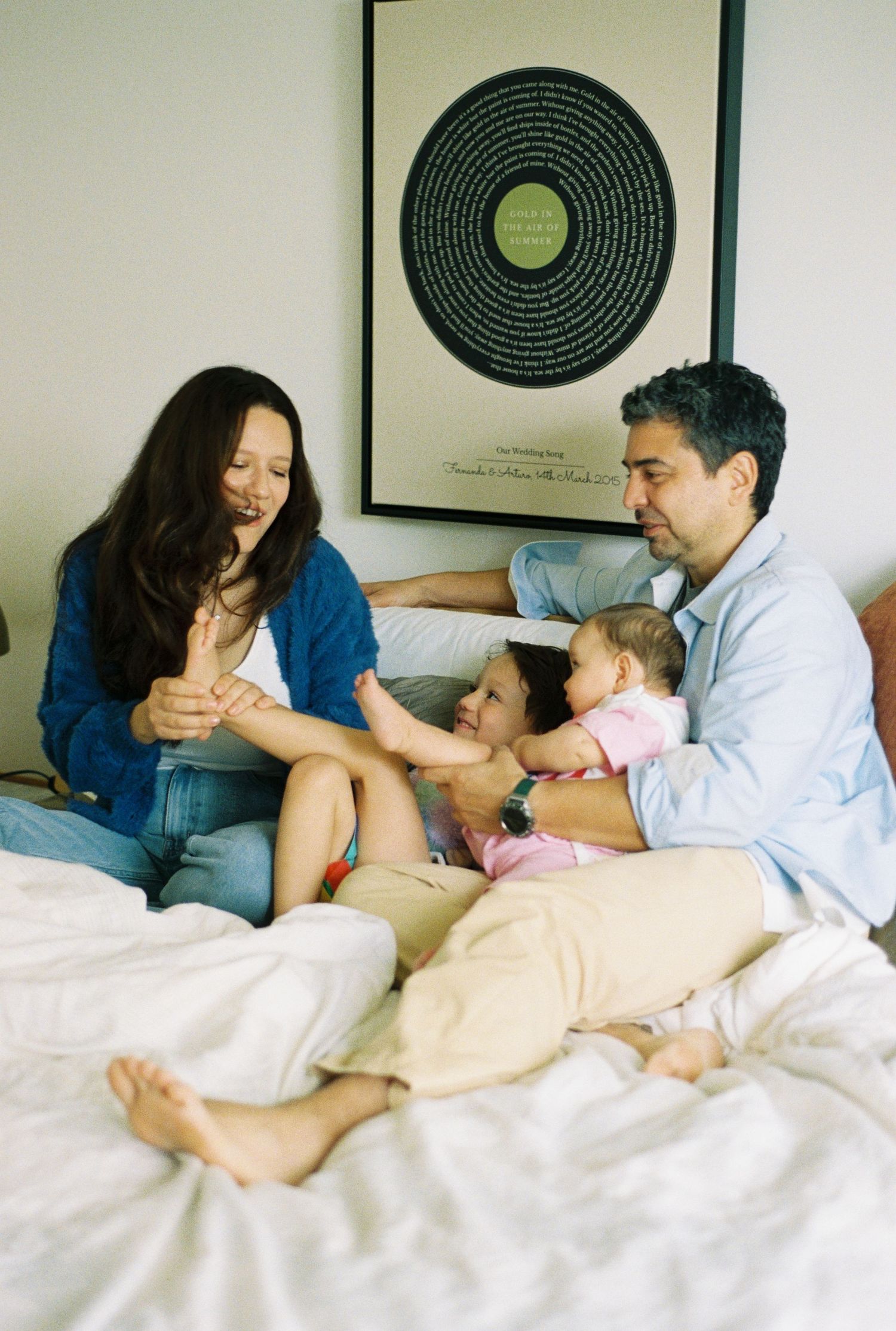 Family of four relaxing on a white bed, parents smiling at two young children, with a vinyl record art print on the wall.
